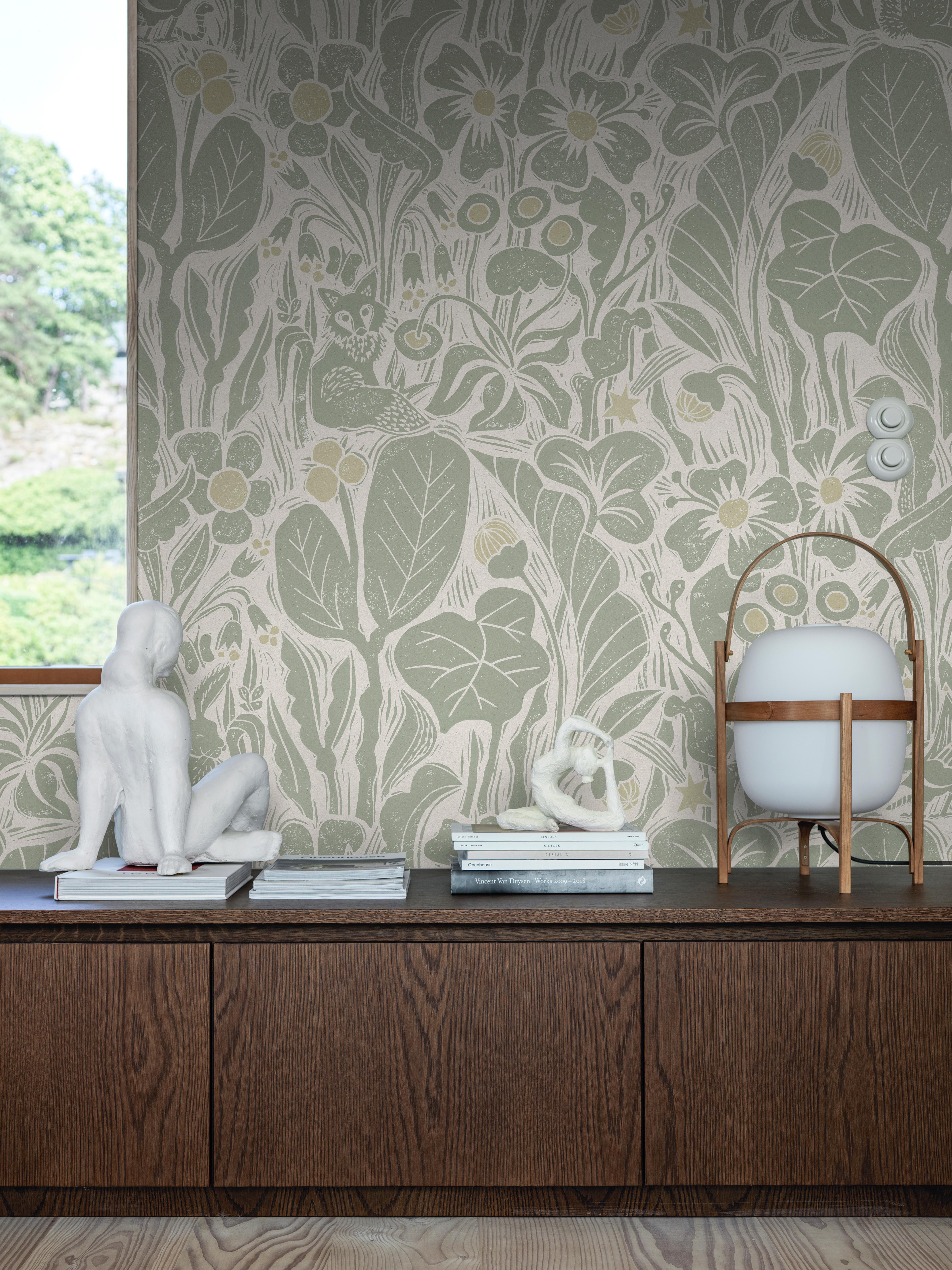 A dark wood credenza with white sculptures and a globe lamp, set against light green wallpaper featuring a nature-inspired woodcut pattern of flowers, leaves, and a fox.