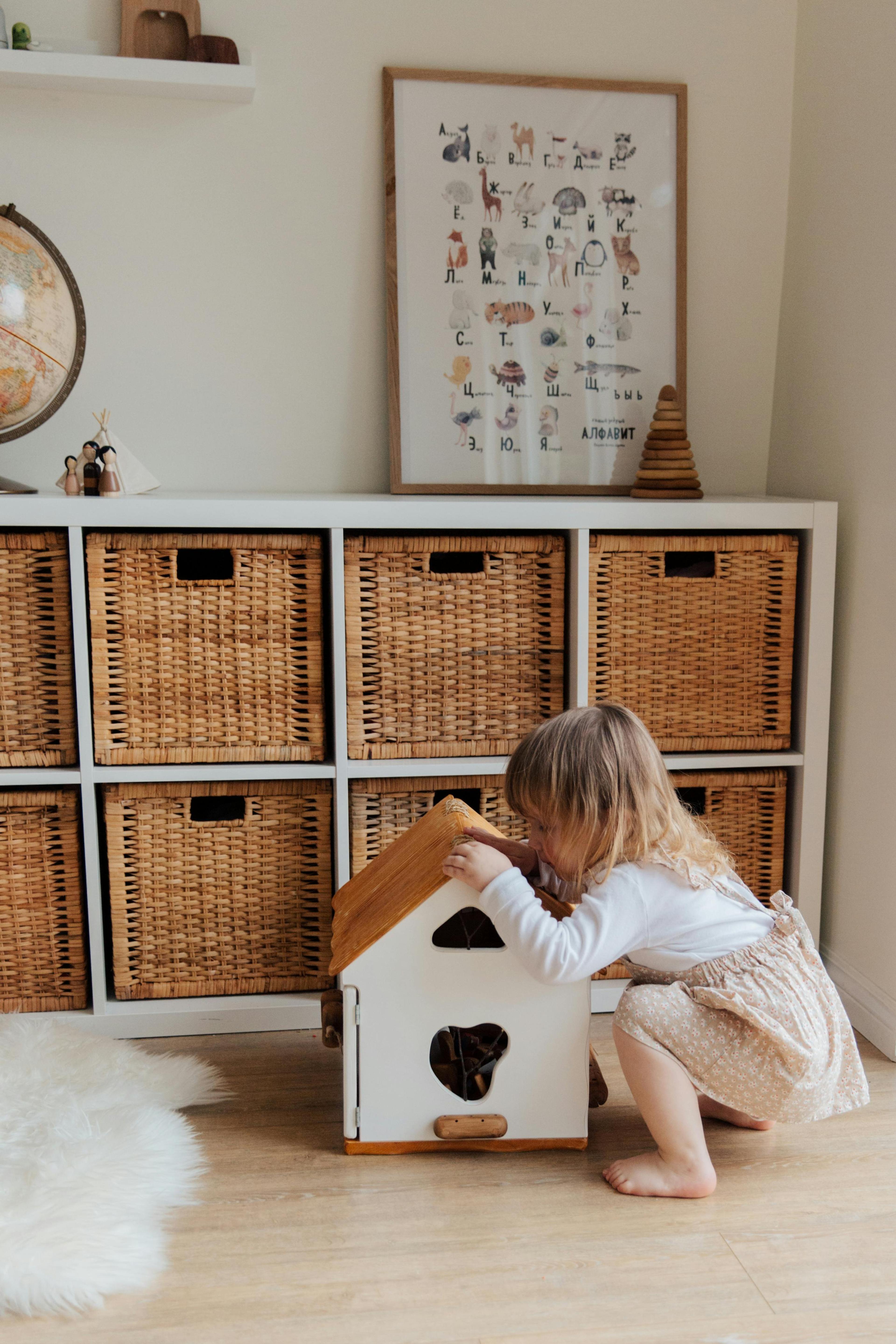 A young child crouches to play with a white wooden toy house in a room with a cubby shelf full of wicker baskets.