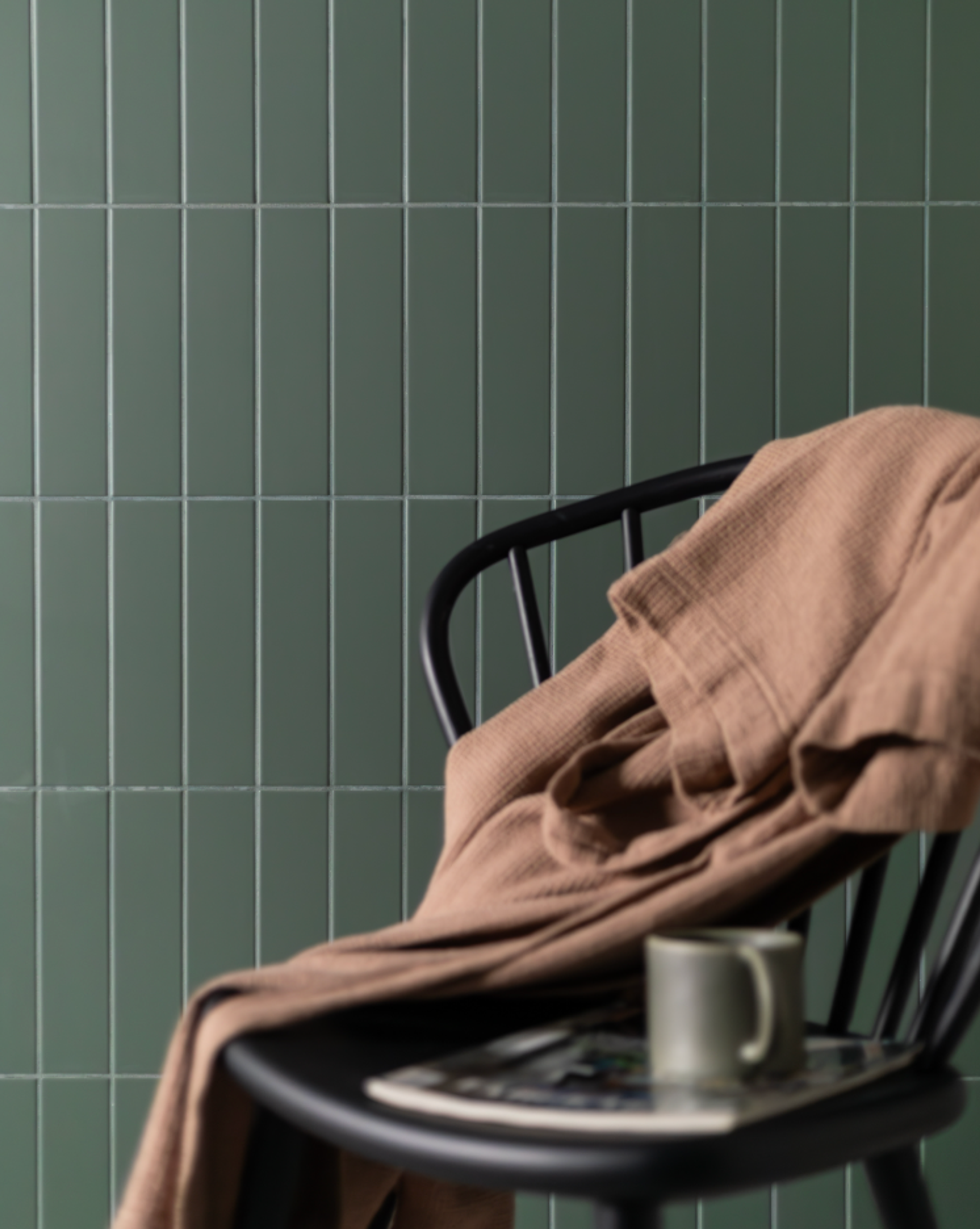 A black chair with a brown blanket, magazine, and green mug in front of a vertical dark green tiled wall.