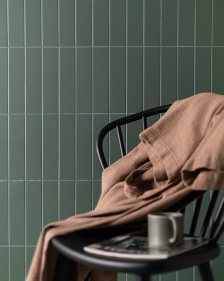 A black chair draped with a brown blanket, a magazine, and a mug sits in front of a wall of vertically stacked green tiles.