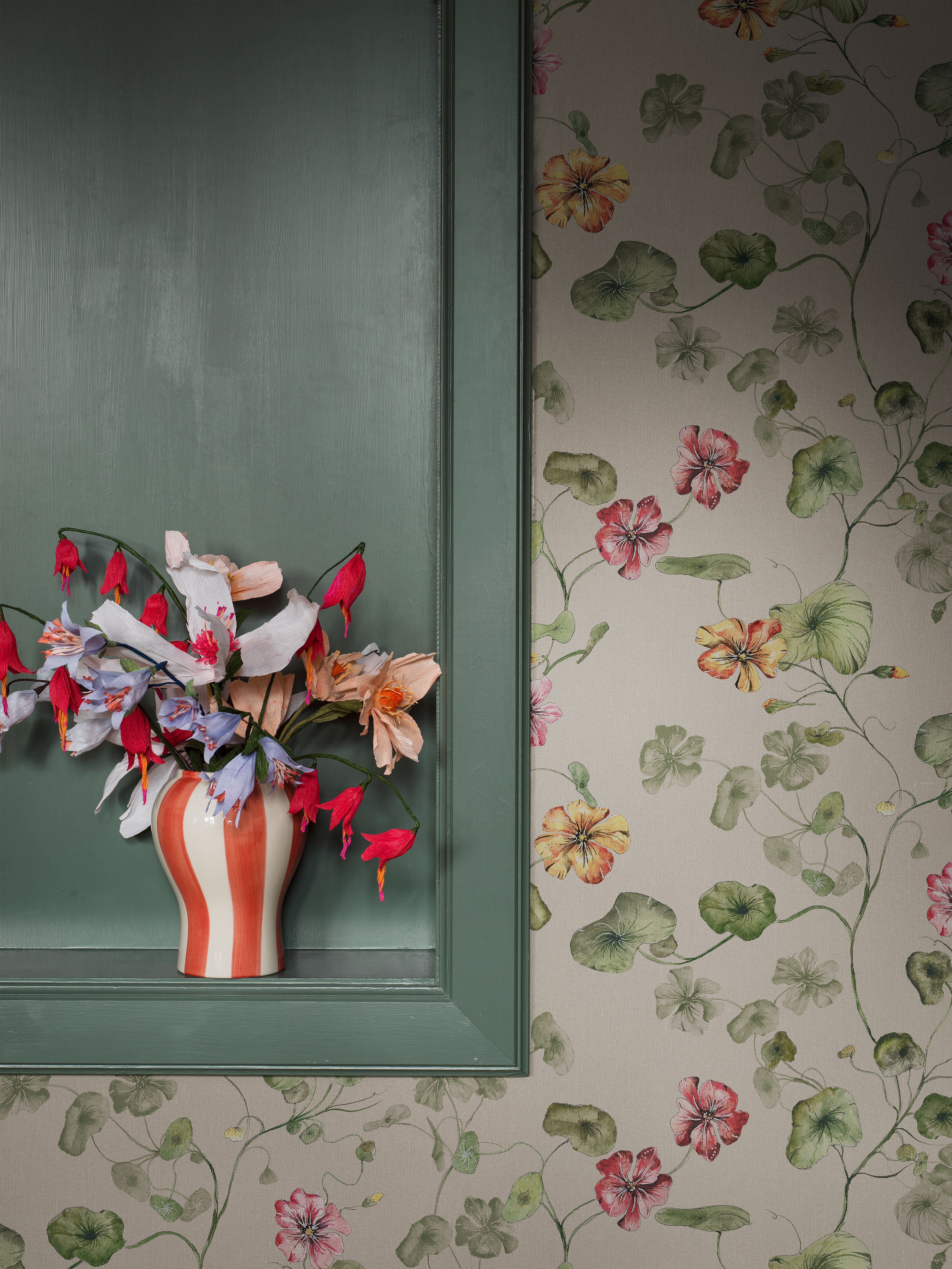 Colorful flowers in a red and white striped vase on a green recessed shelf, against floral wallpaper with green leaves and colorful blossoms.