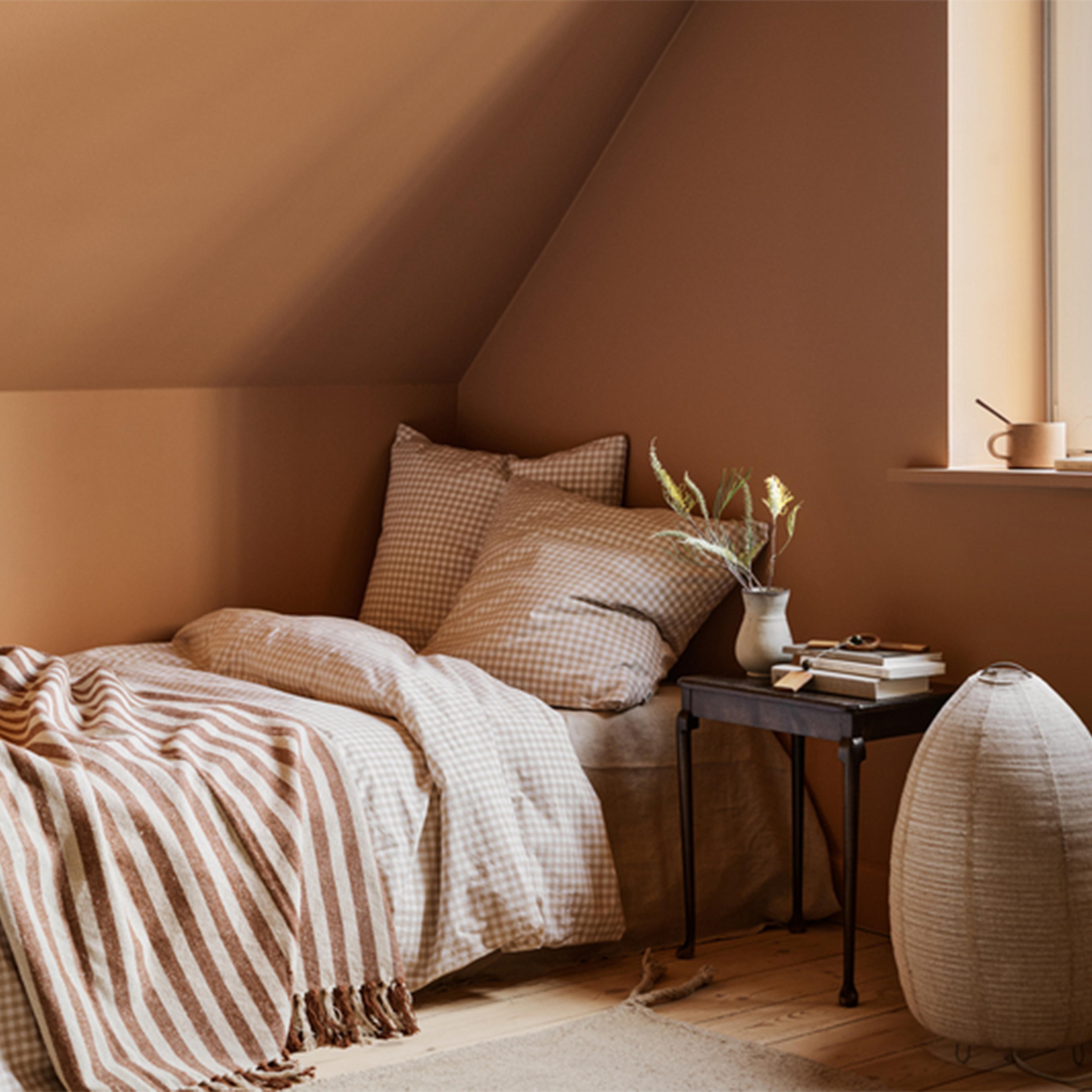 A cozy attic bedroom with warm brown walls, a bed with gingham bedding and a striped throw, a side table with a vase and books, and a large woven floor lamp.