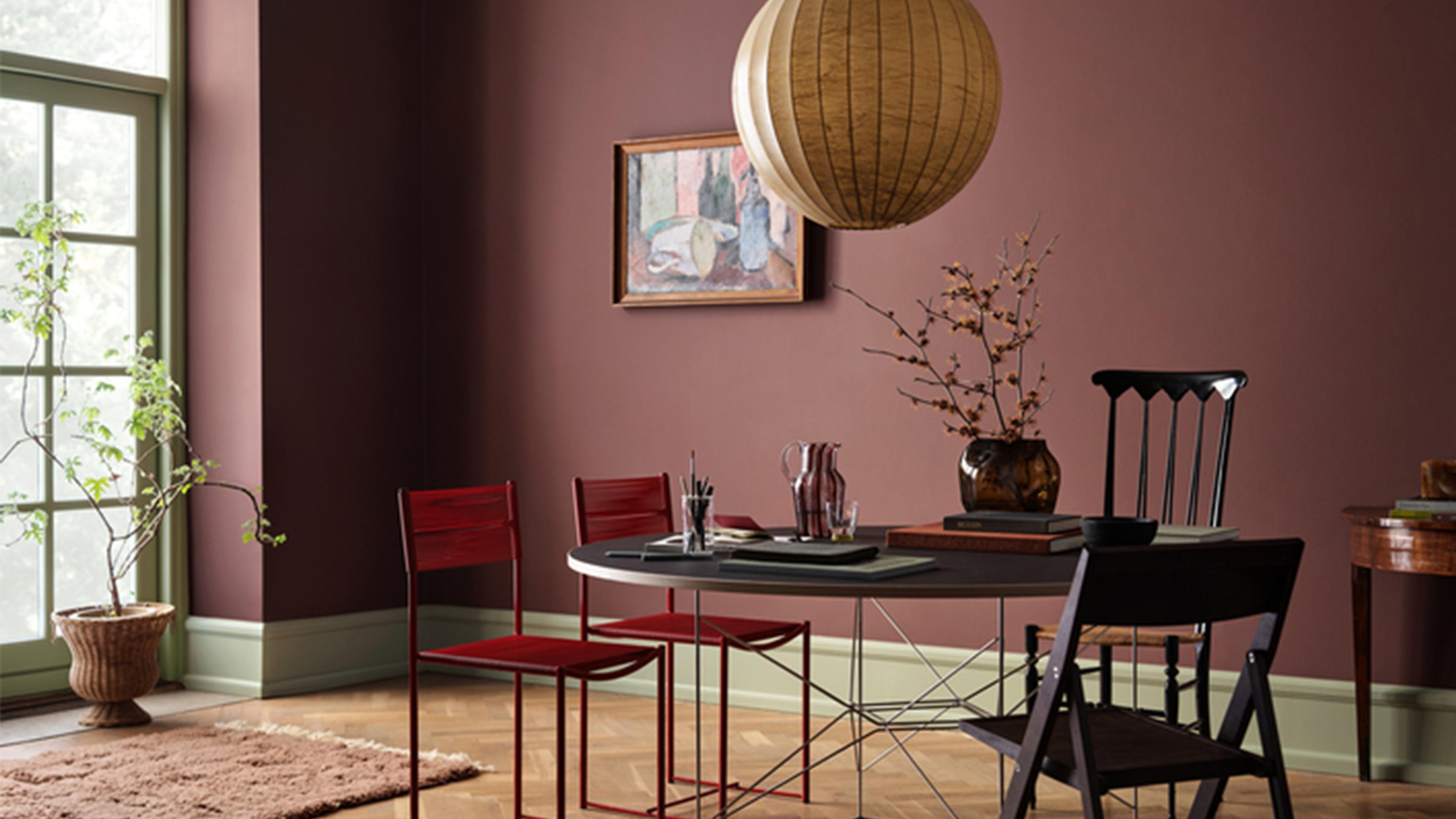 A stylish room with deep plum walls, green trim, a round dining table with mixed chairs, a large paper lantern, and a potted plant by a window.