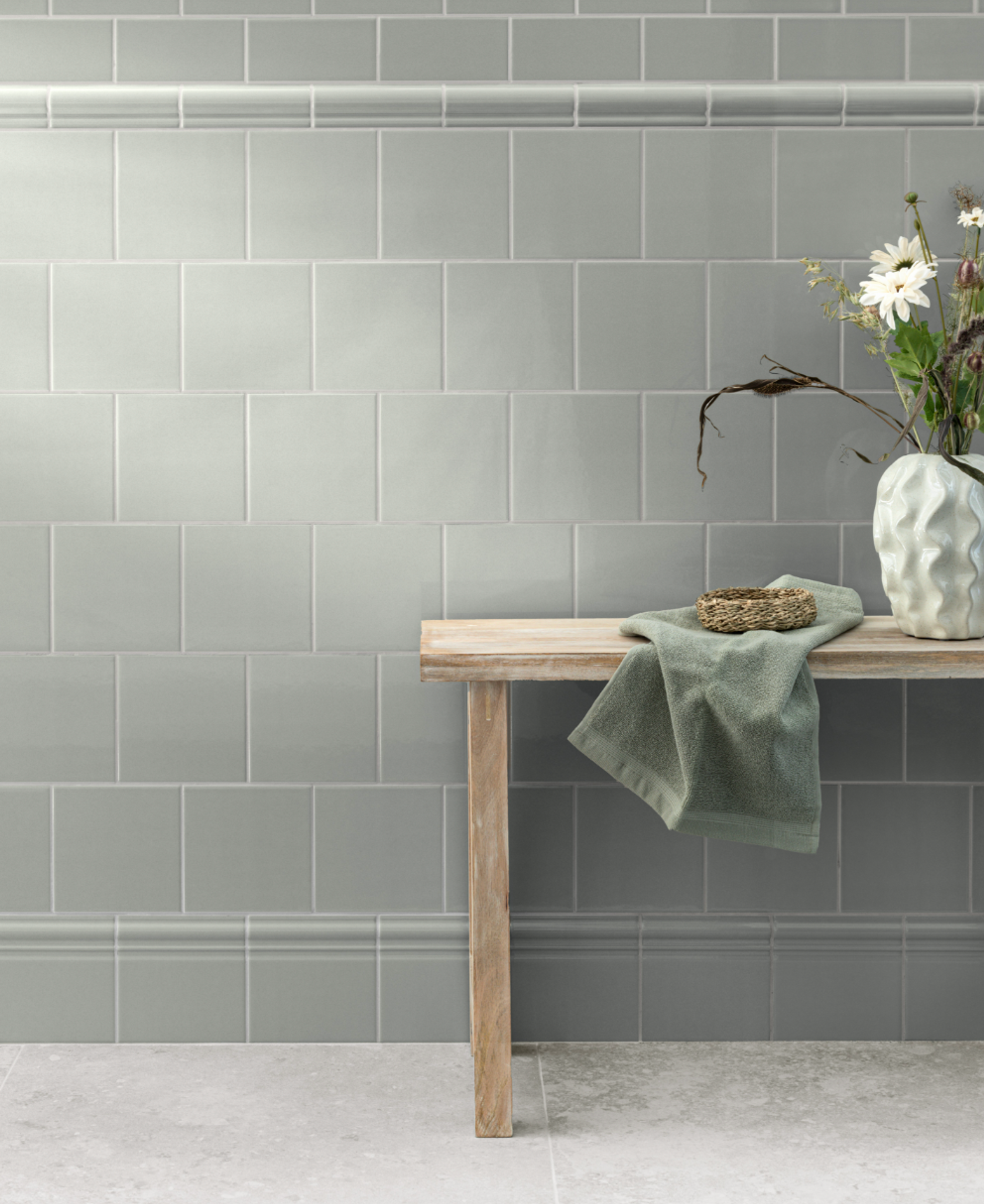 A light gray-green tiled wall with decorative trim, a wooden bench holding a green towel, a small woven basket, and a vase of white daisies.
