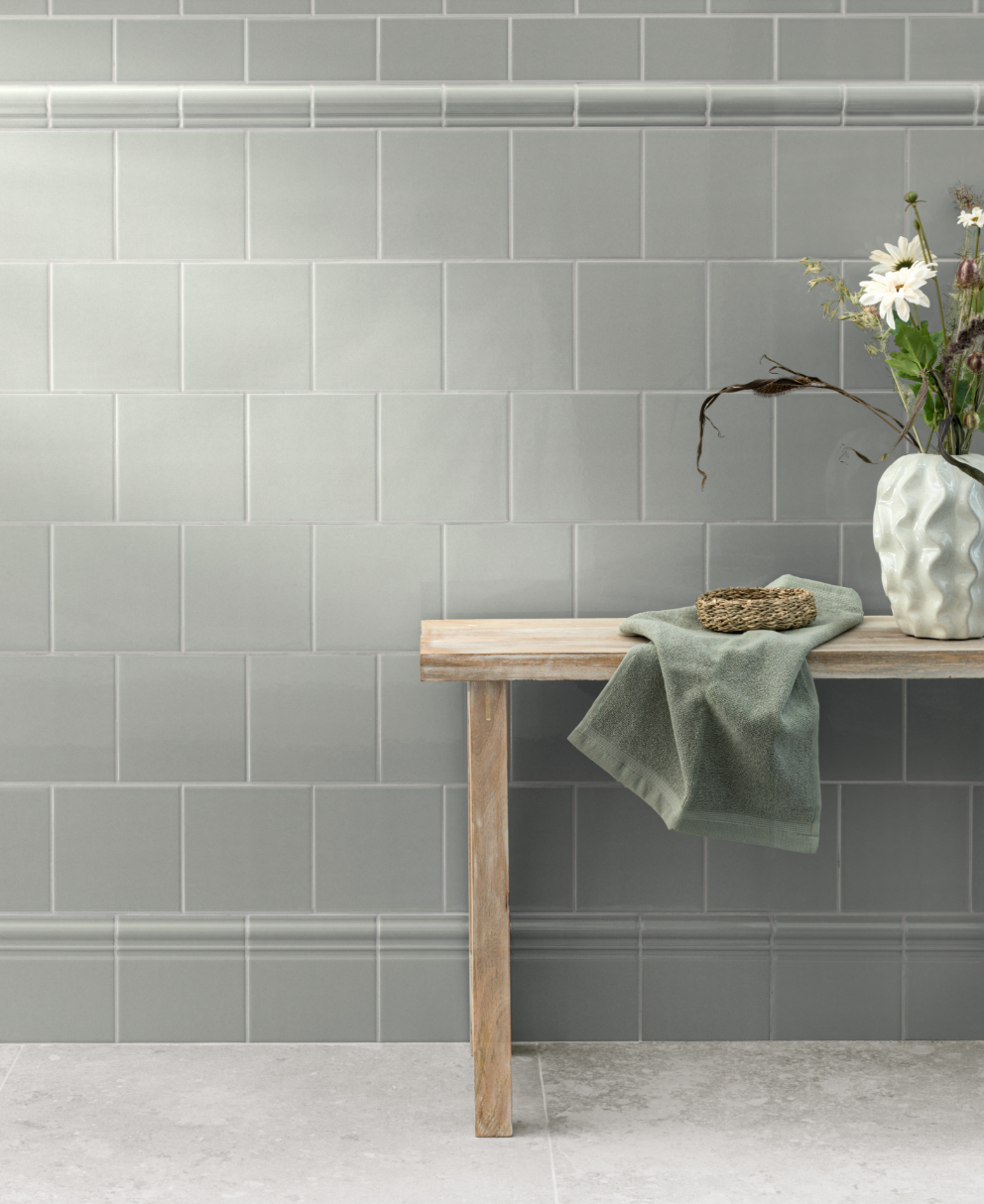 A light gray-green tiled wall with decorative trim, a wooden bench holding a green towel, a small woven basket, and a vase of white daisies.