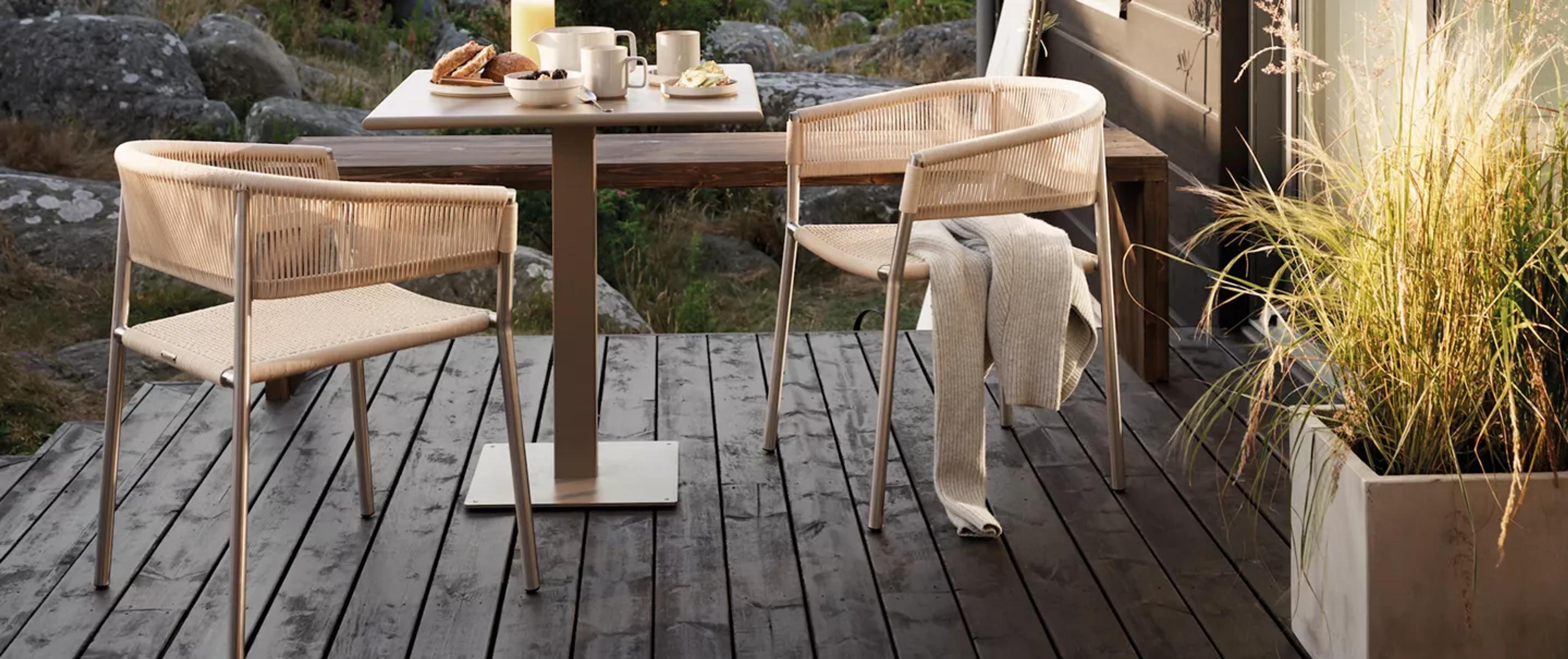 Outdoor breakfast on a wooden deck with two woven chairs and a table, next to natural rocks and tall grasses.