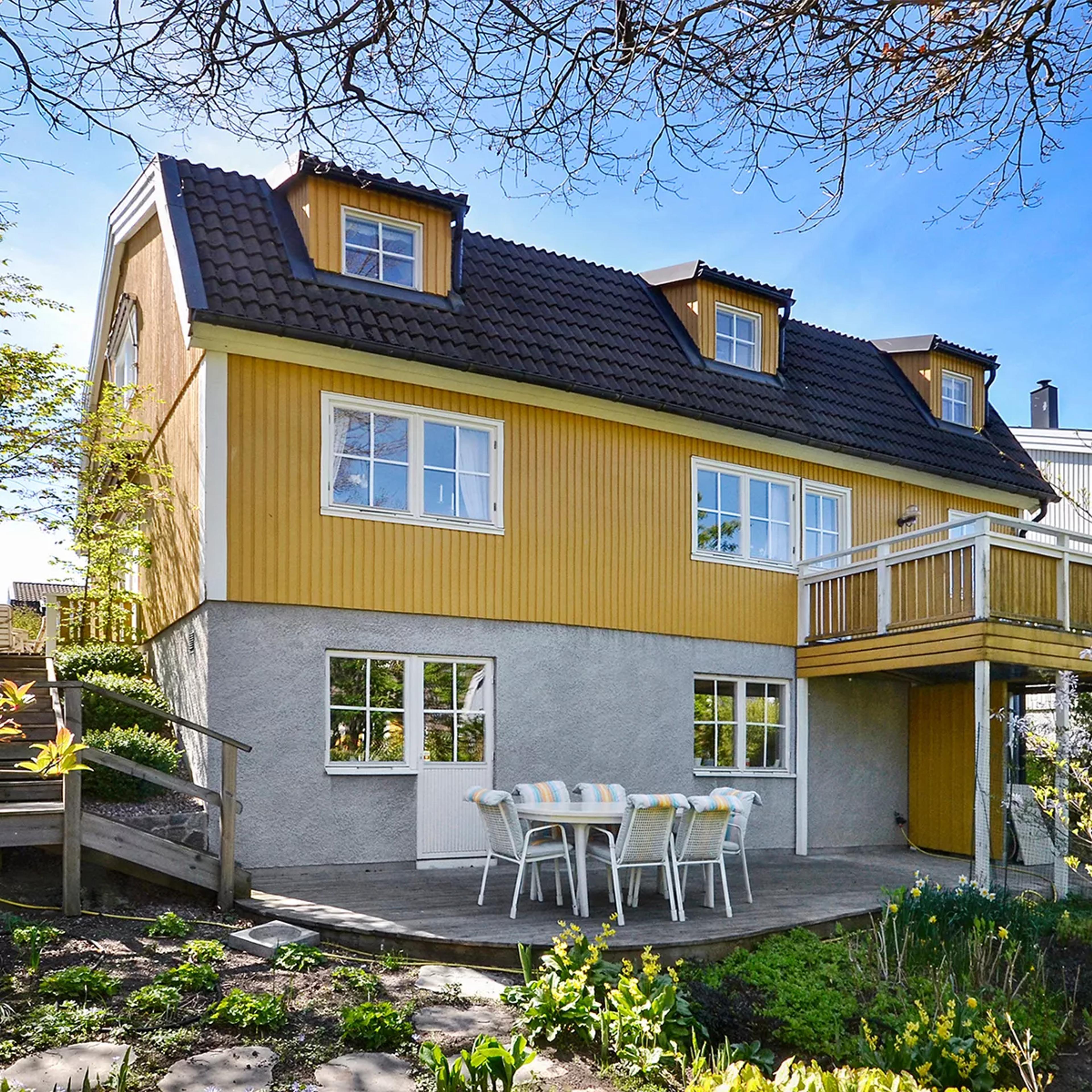 A yellow house with a grey stucco base, dark roof, a balcony, and a patio with outdoor furniture in a garden.