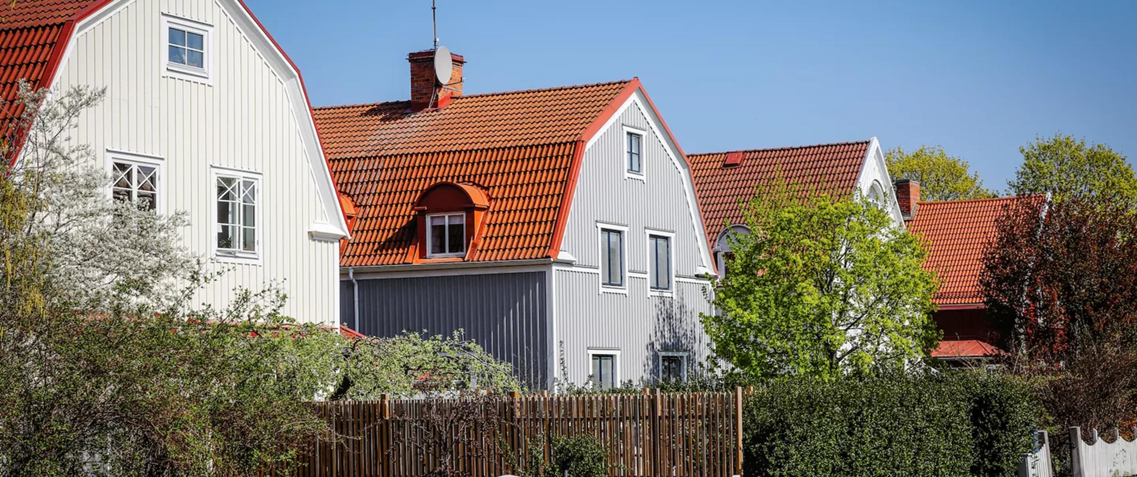 A row of homes with light-colored vertical siding and red tiled roofs, behind a wooden fence and green trees.