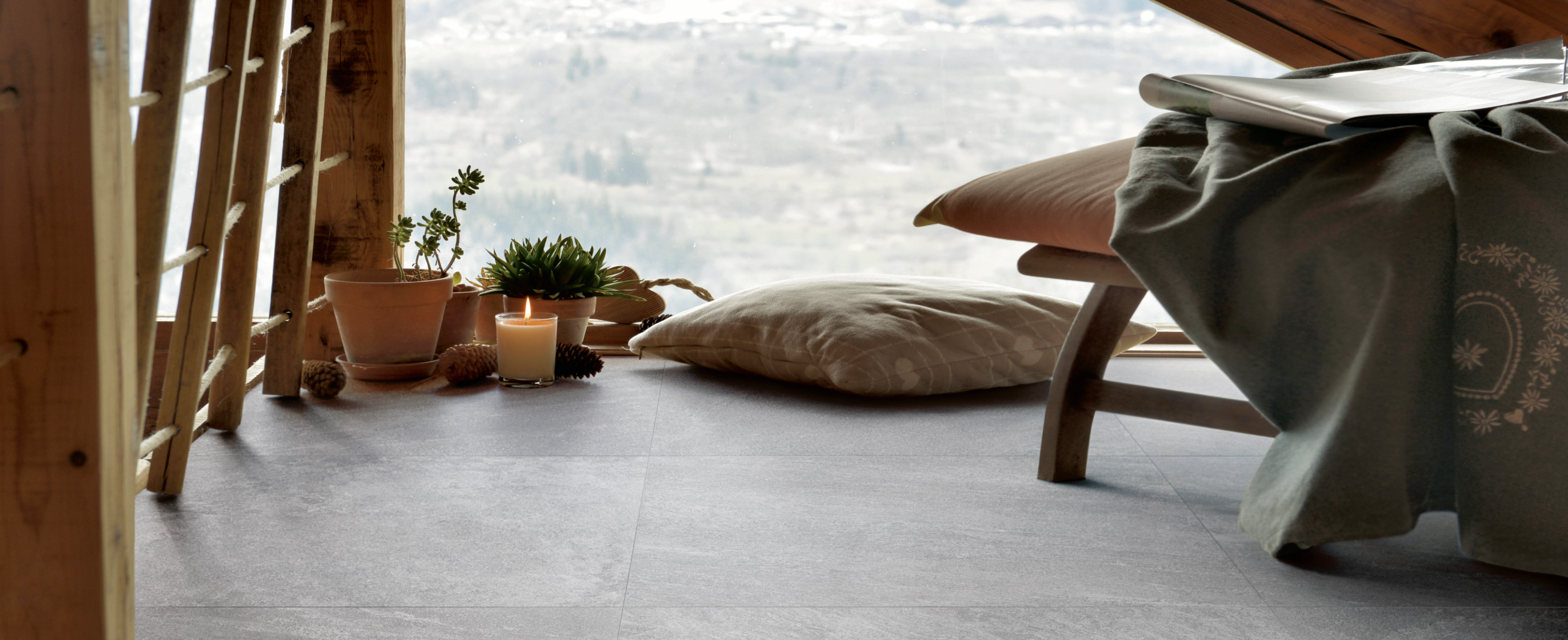 A cozy indoor scene with a dark grey floor, potted plants, a lit candle, cushions, and a blanket-draped stool, all facing a large window with a hazy mountain view.