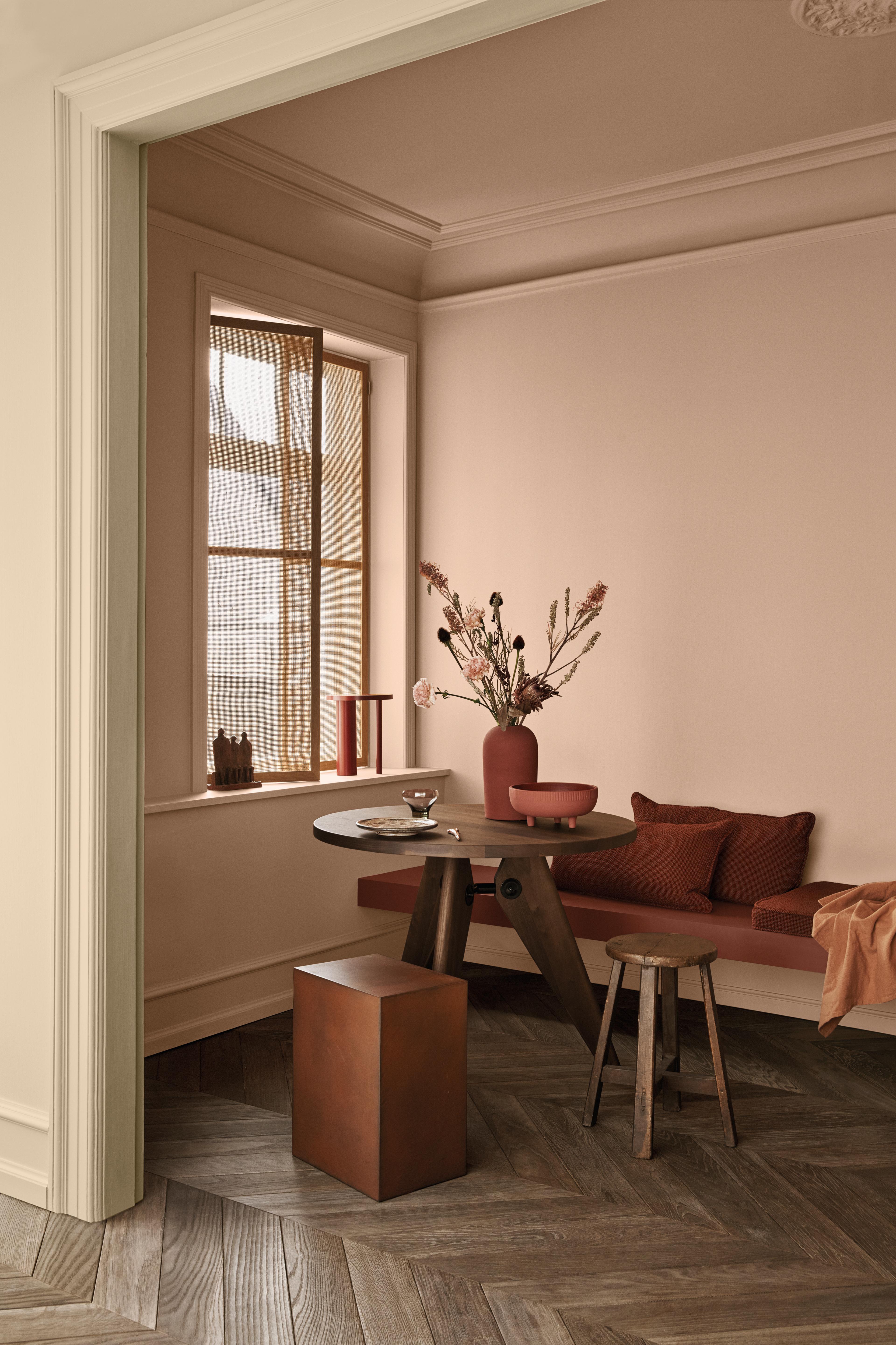 A warm dining nook with terracotta pink walls, a round wooden table, a built-in bench with rust-colored cushions, and a herringbone wooden floor.