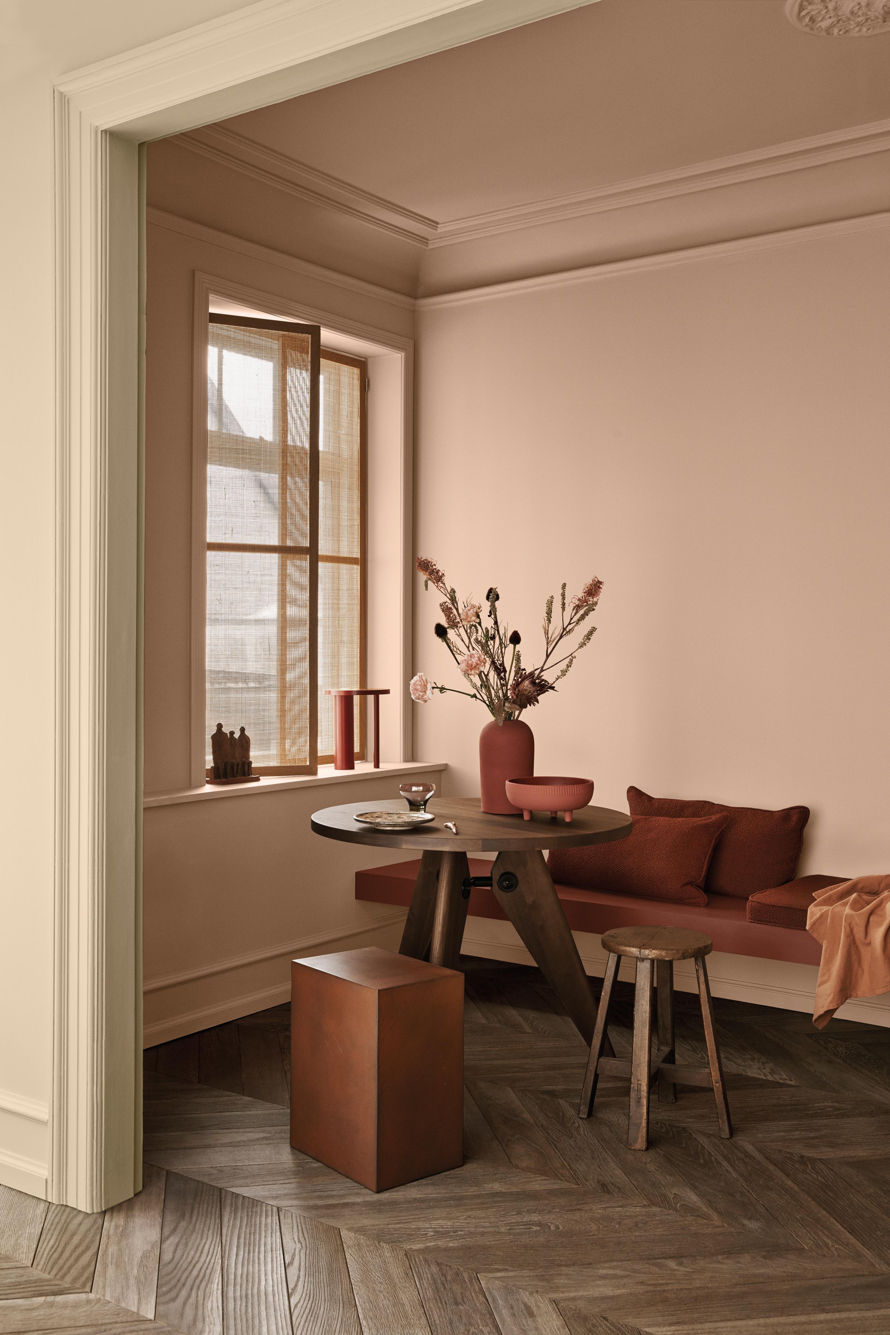 A warm dining nook with terracotta pink walls, a round wooden table, a built-in bench with rust-colored cushions, and a herringbone wooden floor.