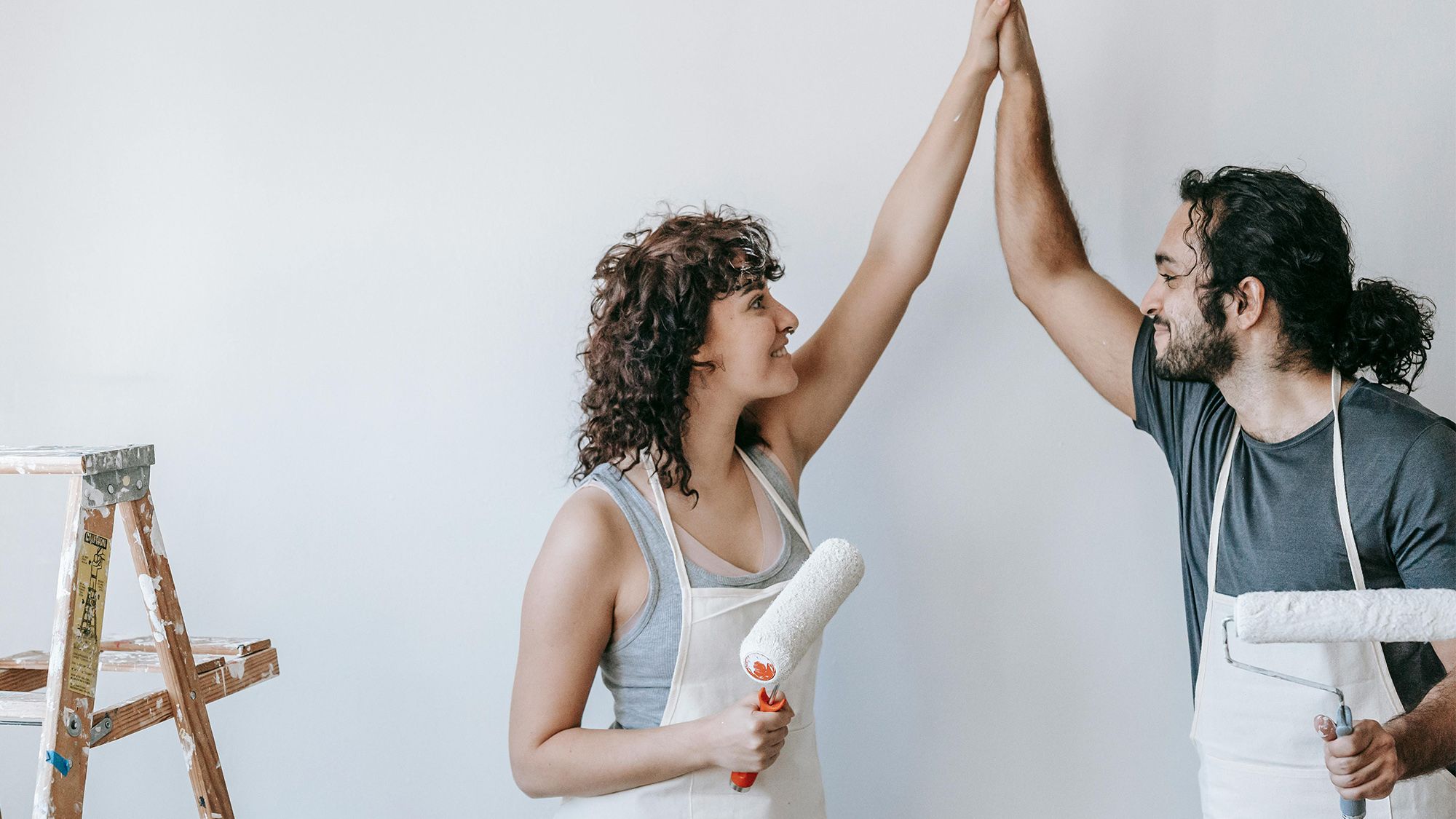 A man and woman in aprons, holding paint rollers, high-fiving.