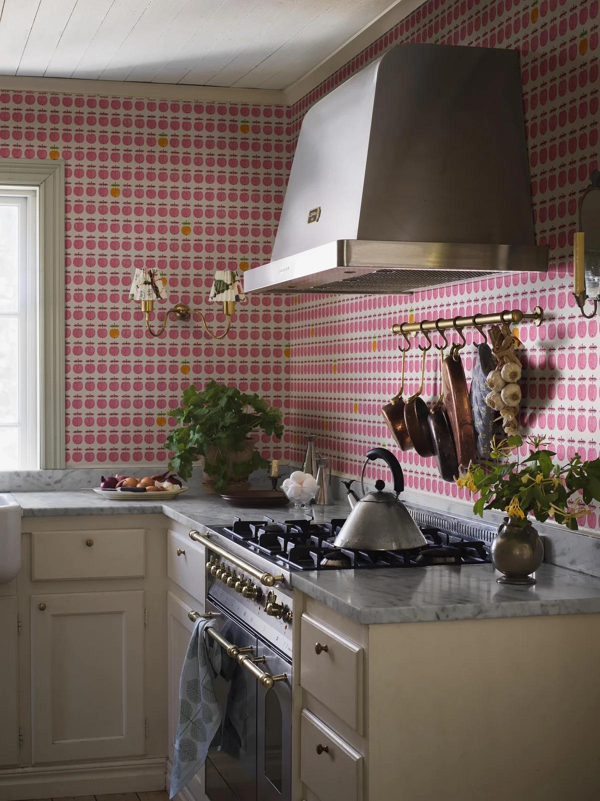 A kitchen with pink and yellow apple-patterned wallpaper, a large stainless steel range hood, a professional stove with brass accents, and marble countertops.