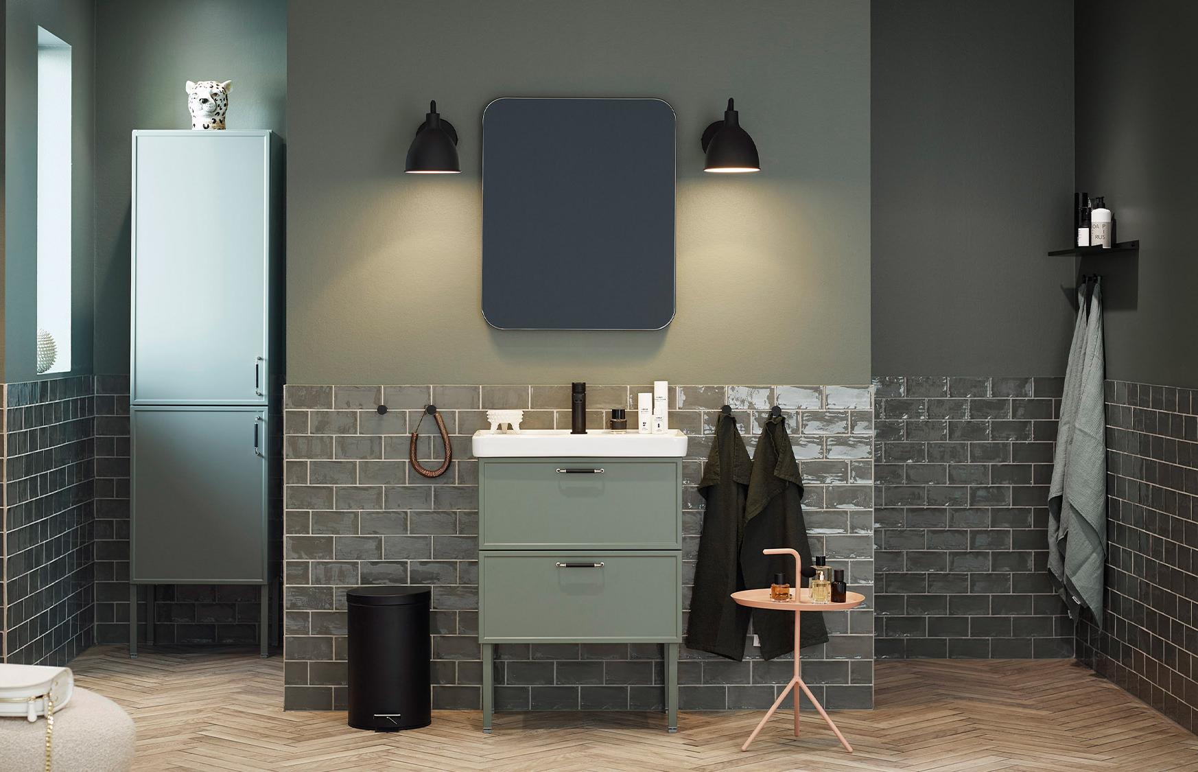 Stylish modern bathroom with olive green walls, dark gray subway tiles, a light green vanity with sink, tall cabinet, dark mirror, and herringbone wood floor.