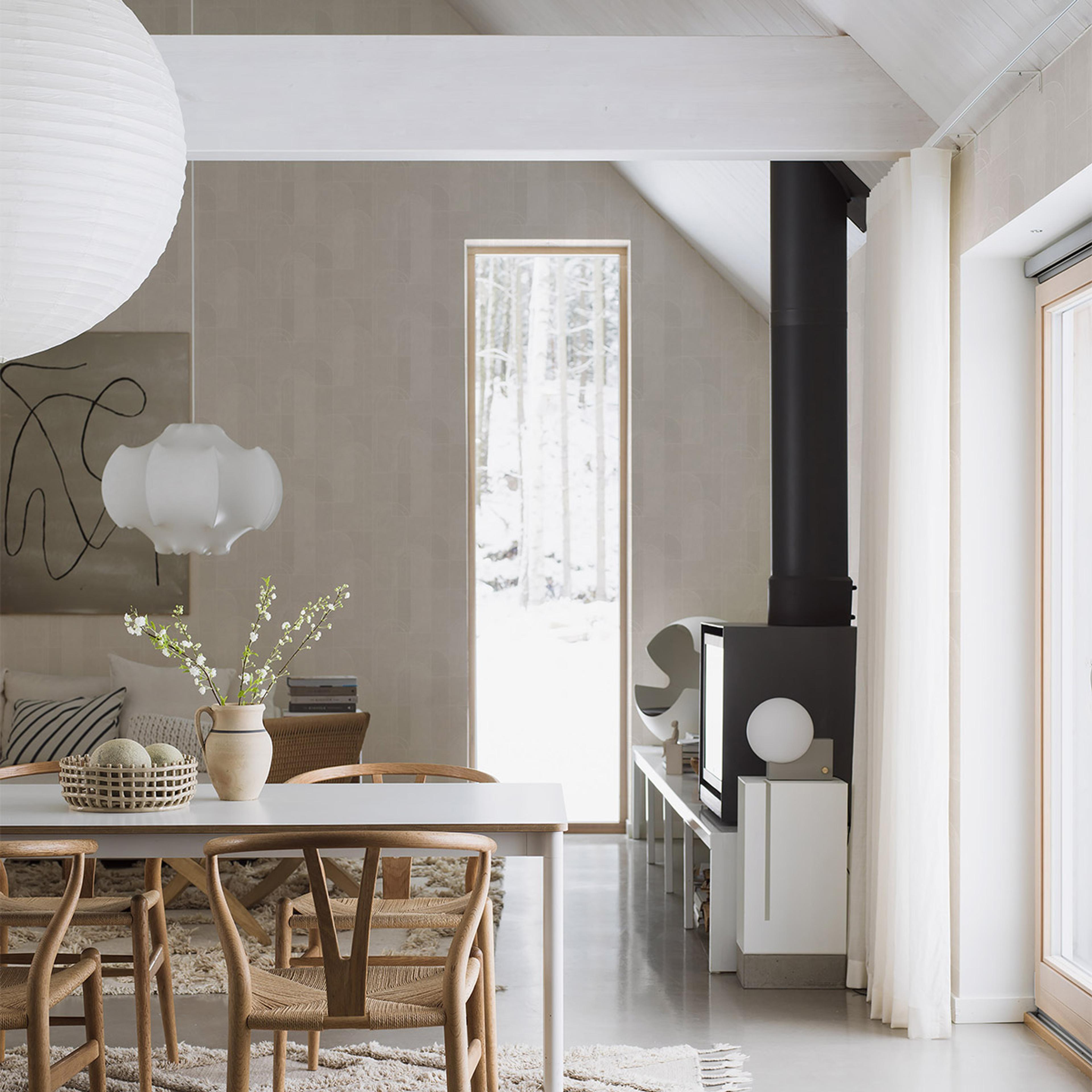 A bright, minimalist interior featuring a dining table with wooden chairs, a large paper lantern, a wood stove, and a tall window overlooking a snowy forest.