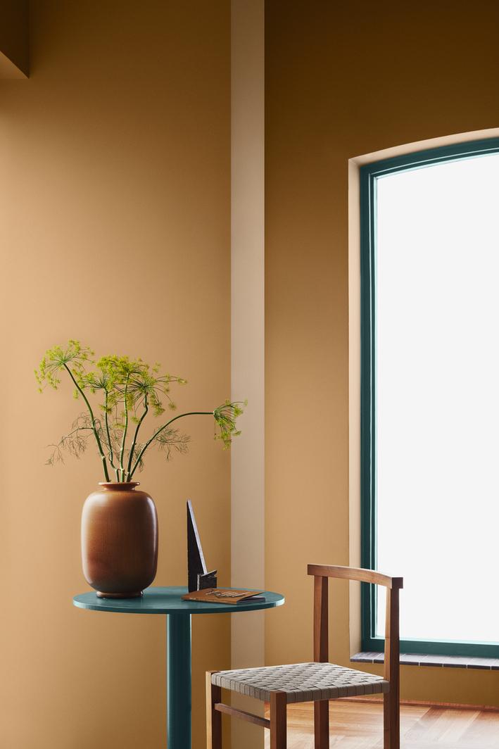 A room with warm brown walls, a white column, a round teal table with a terracotta vase of green plants, a wooden chair, and a large window with teal trim.
