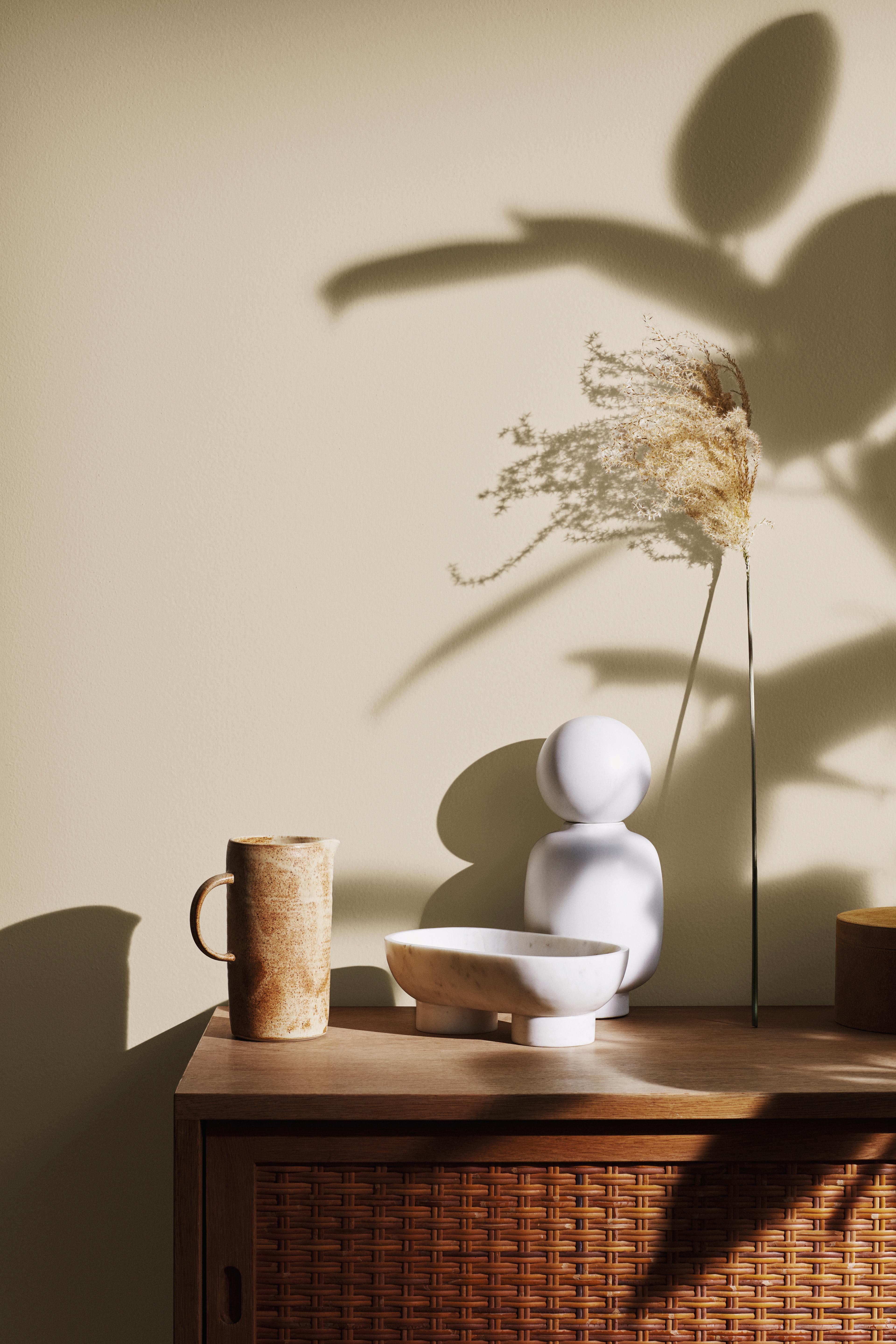 Minimalist still life of a ceramic jug, marble bowl, white sculpture, and dried grass on a rattan cabinet, with plant shadows on a beige wall.