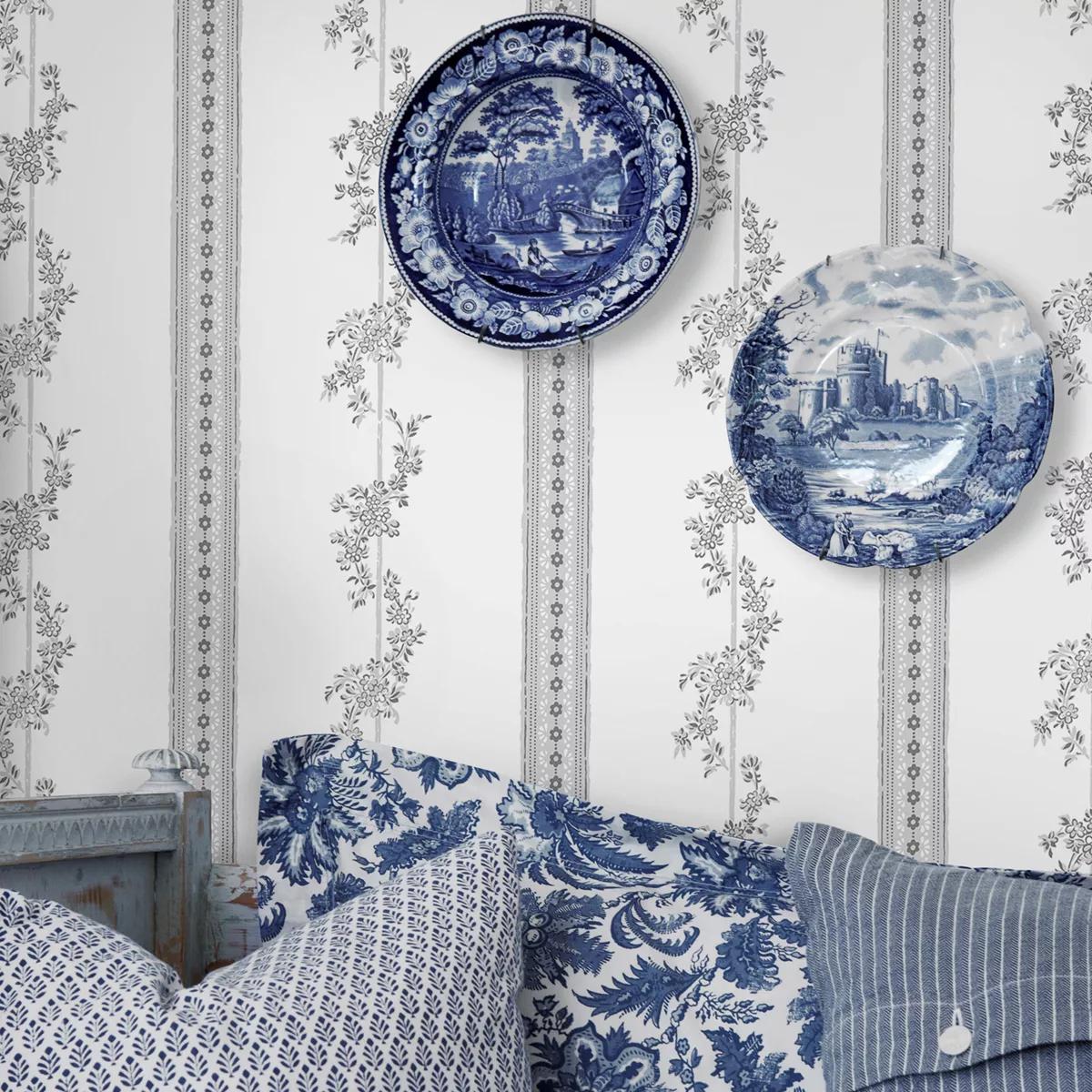 White wallpaper with grey floral vines and narrow patterned stripes, two blue and white decorative plates, and blue patterned pillows.