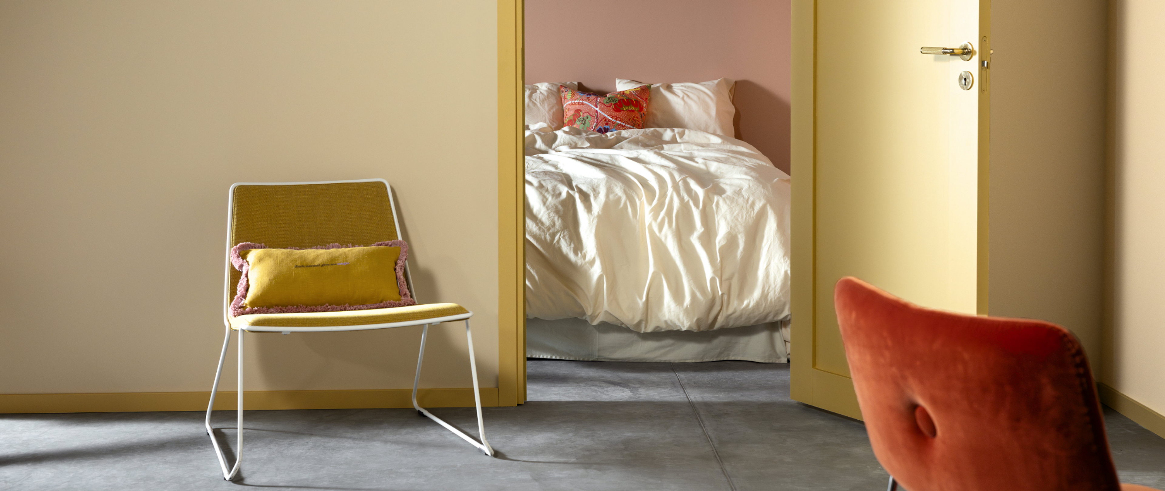 A yellow doorway leads into a pink bedroom with a bed, with a yellow chair and an orange chair in the foreground.