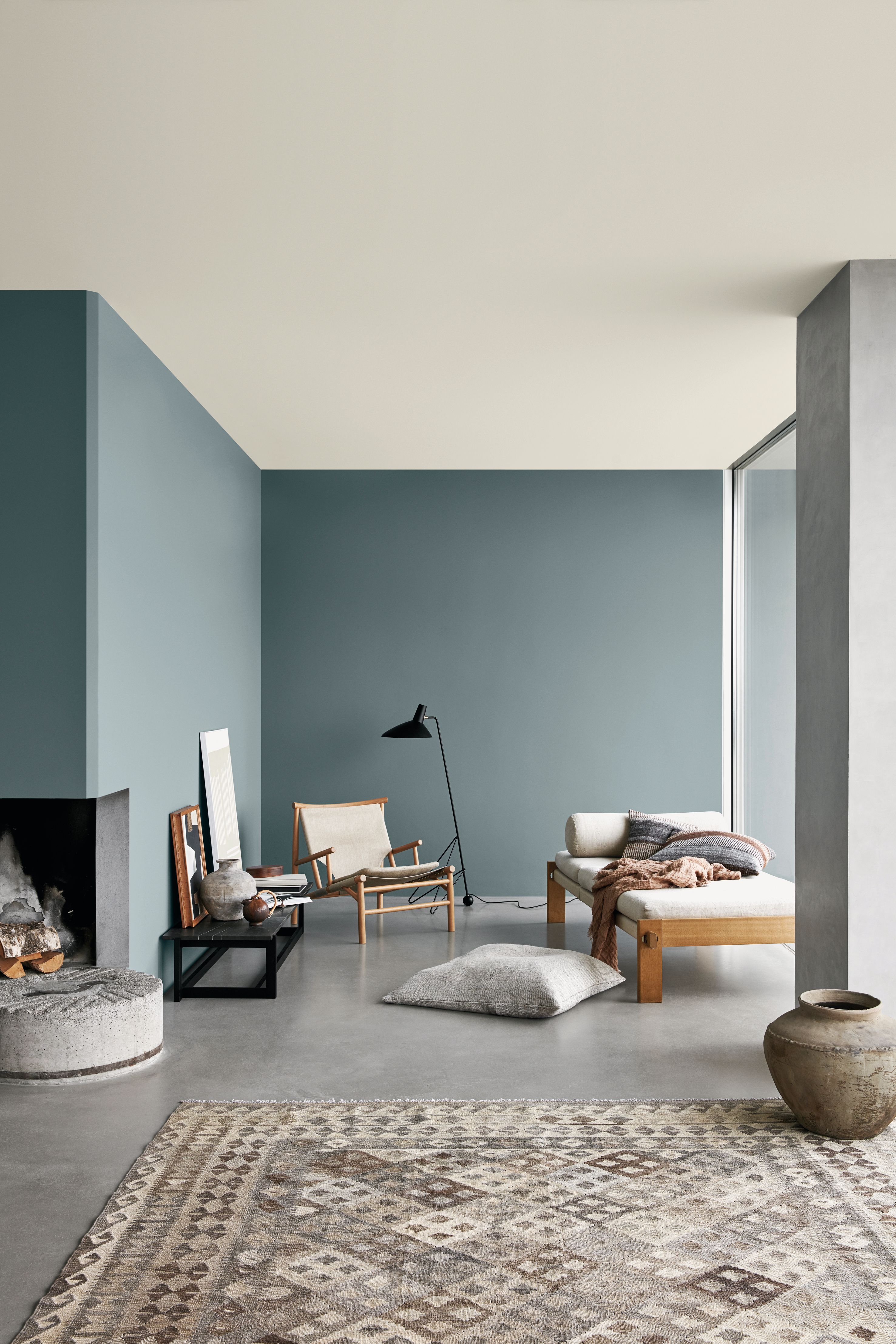A modern living room with muted teal walls, a light ceiling, a concrete floor with a patterned rug, a fireplace, and minimalist wooden furniture.