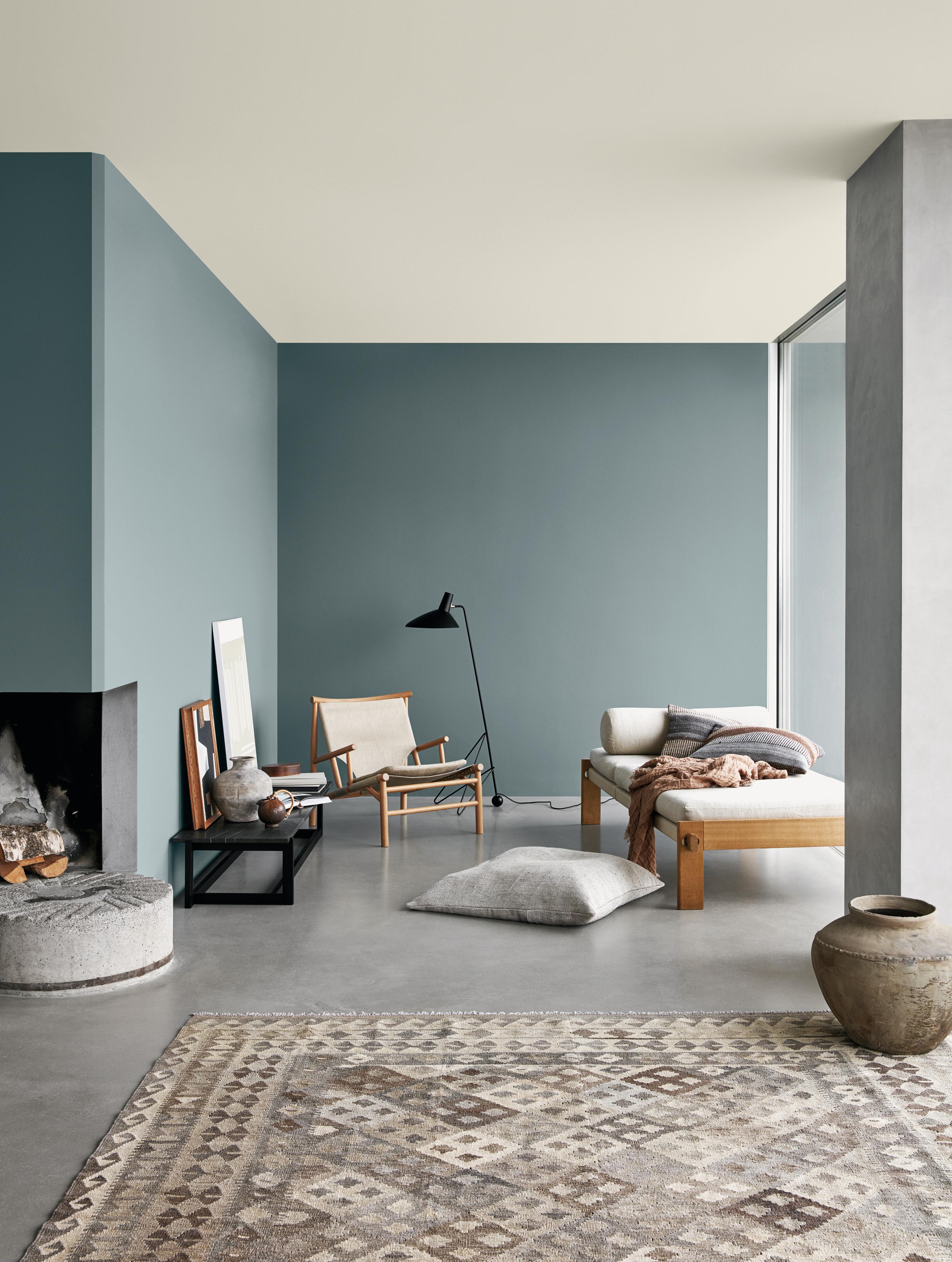 A modern living room with muted teal walls, a light ceiling, a concrete floor with a patterned rug, a fireplace, and minimalist wooden furniture.