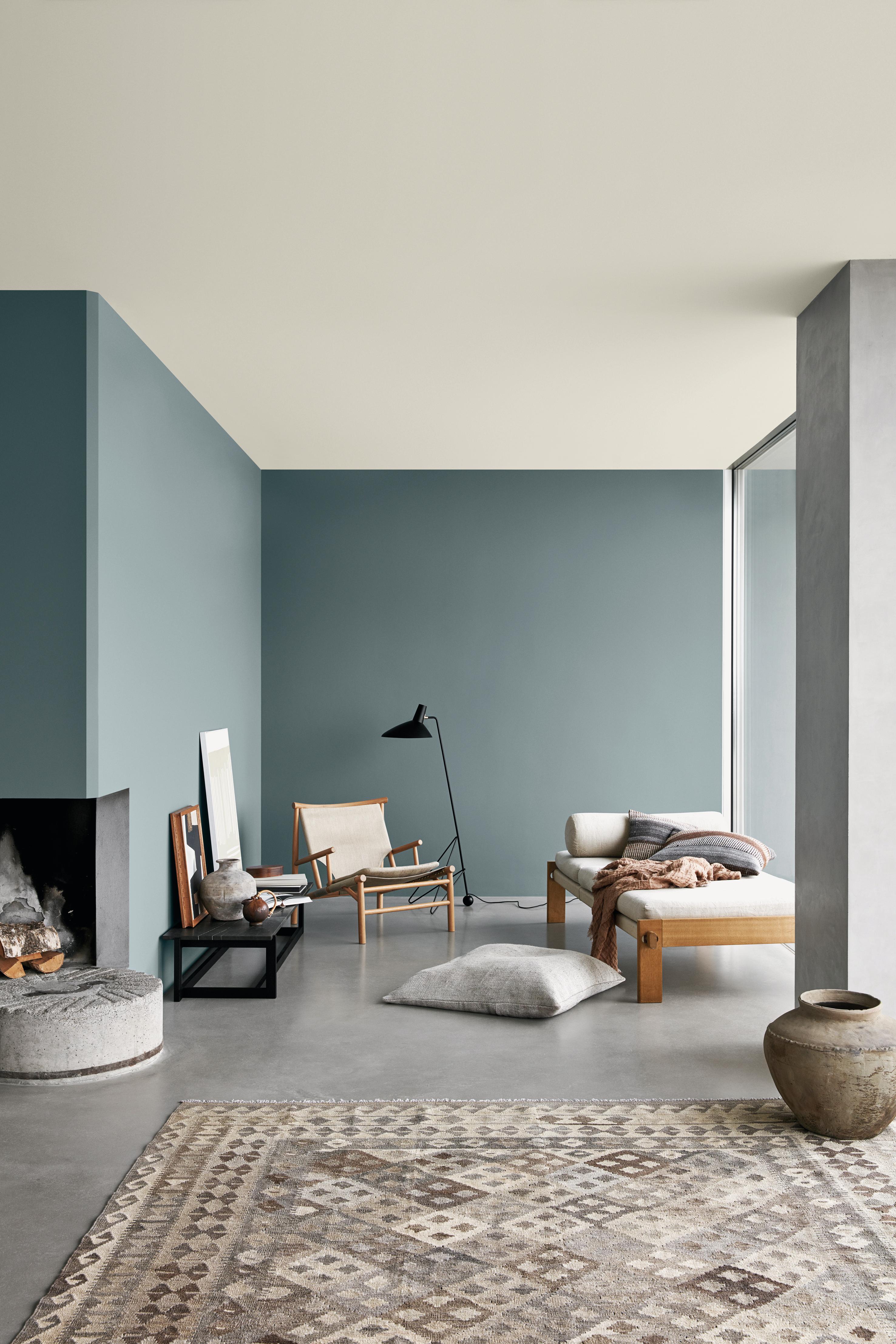 A modern living room with muted teal walls, a light ceiling, a concrete floor with a patterned rug, a fireplace, and minimalist wooden furniture.