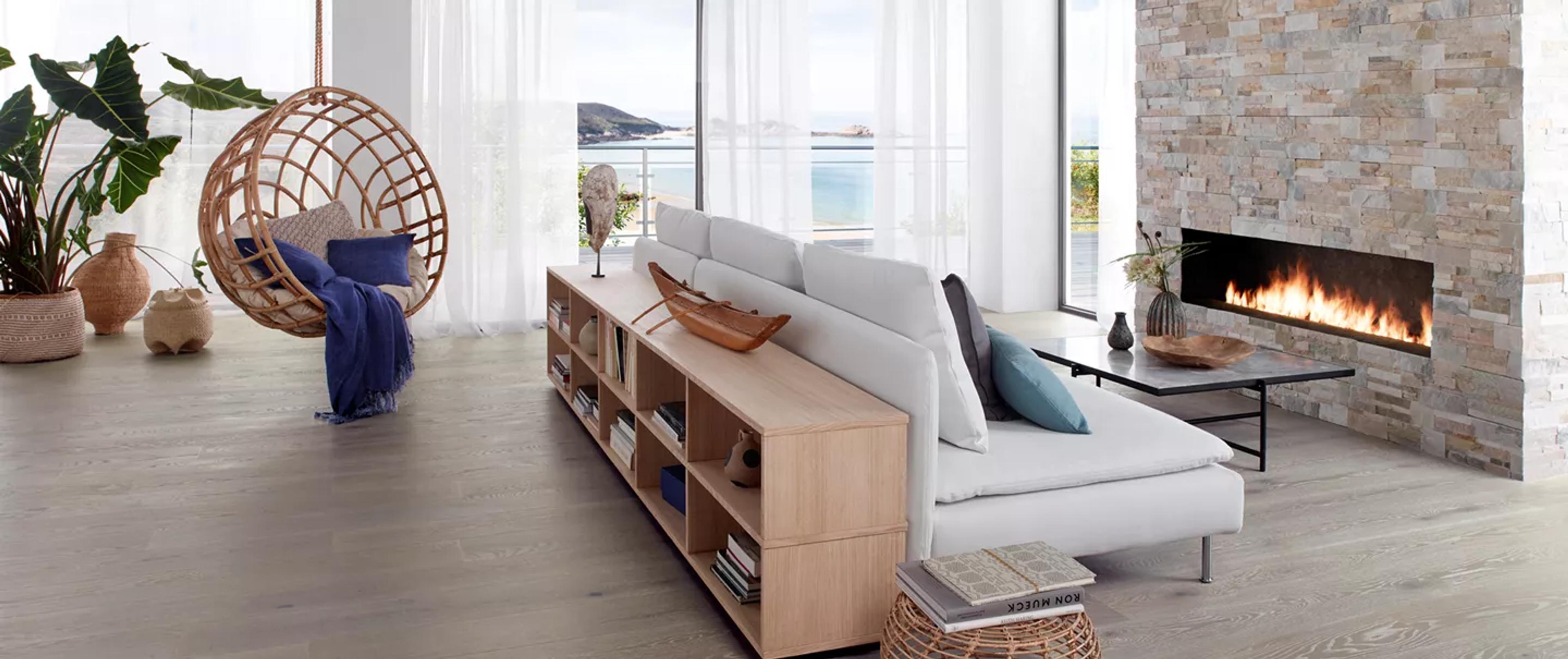 Modern living room with light wood floors, a hanging wicker chair, a white sofa backed by a wooden bookshelf, a stone fireplace with flames, and an ocean view.
