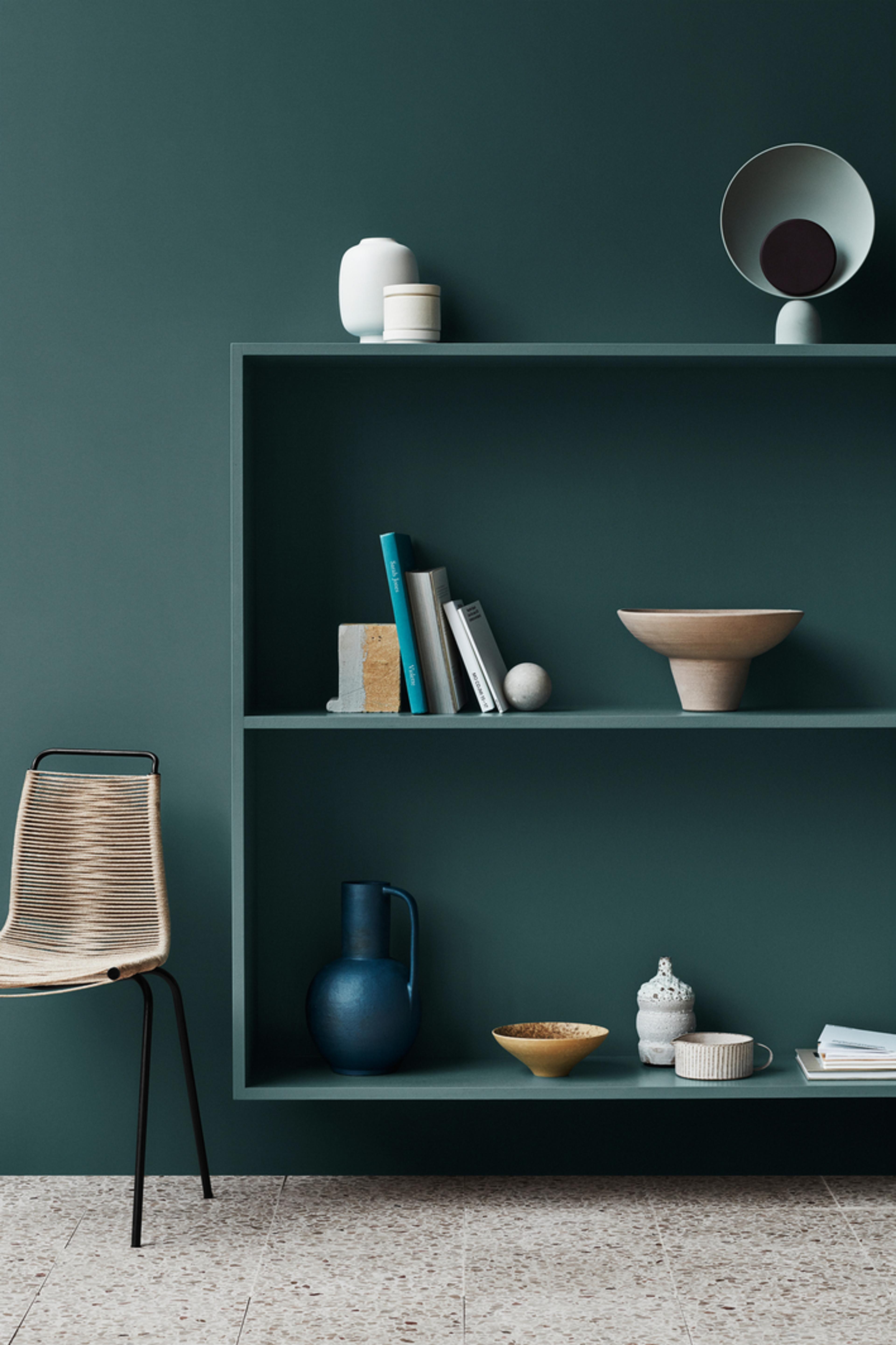 A modern room featuring a deep teal wall with matching built-in shelves holding minimalist decor, a woven chair, and a light terrazzo floor.