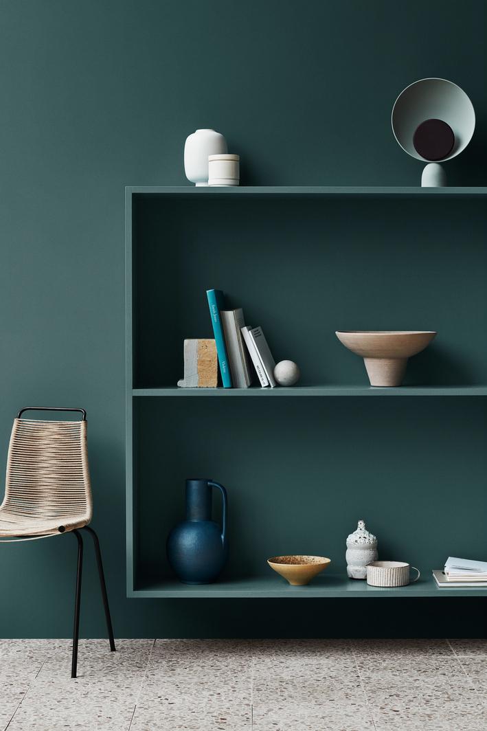 A modern room featuring a deep teal wall with matching built-in shelves holding minimalist decor, a woven chair, and a light terrazzo floor.