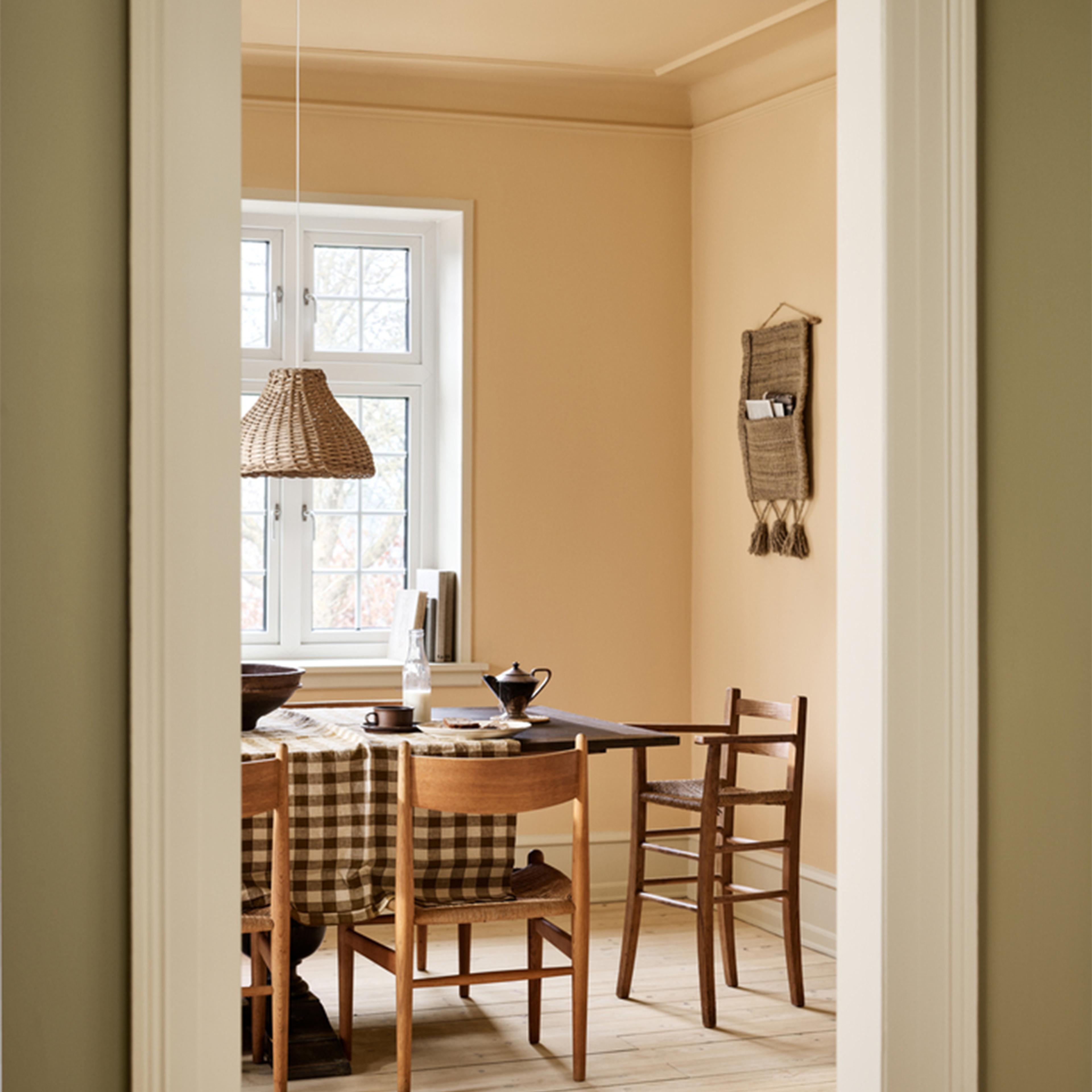 A dining room with peach walls, a wooden table with a checkered tablecloth, and a wicker lamp, viewed through a doorway.