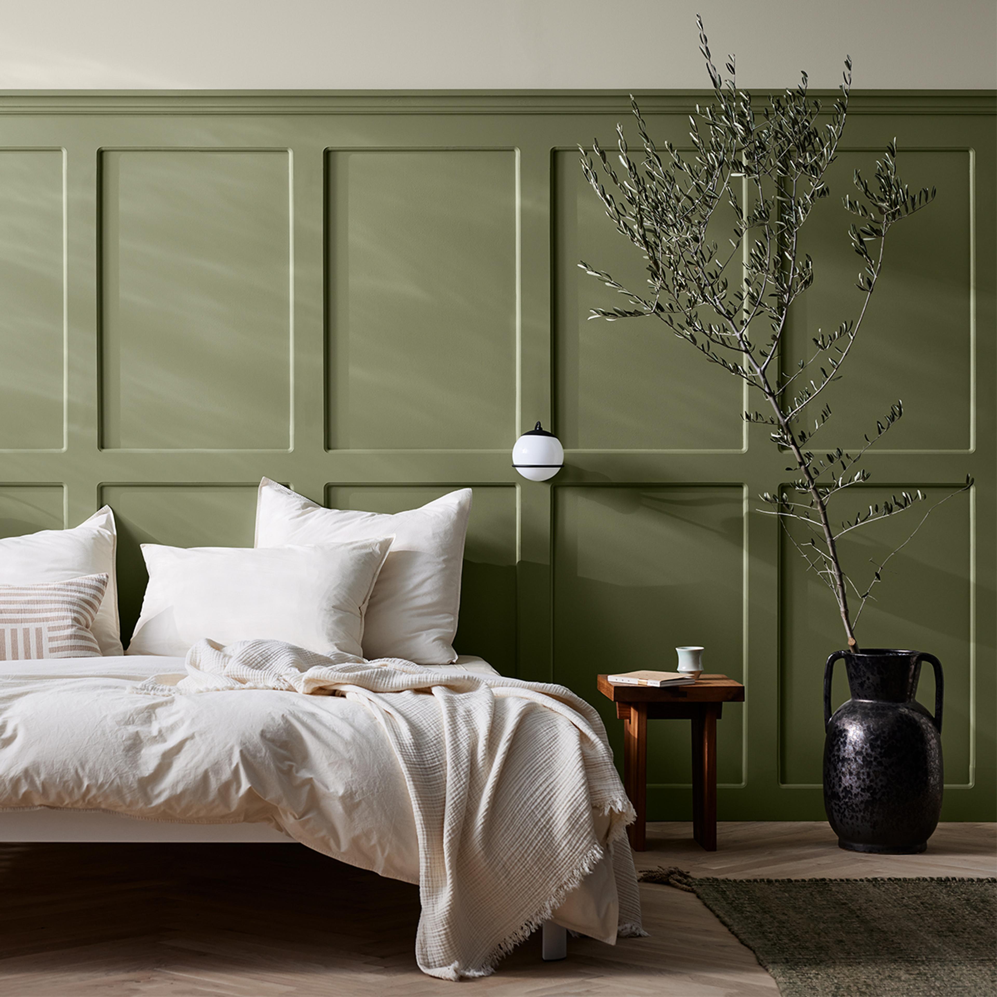 A bedroom with a sage green paneled wall, a bed with white bedding, and an olive branch in a dark vase.