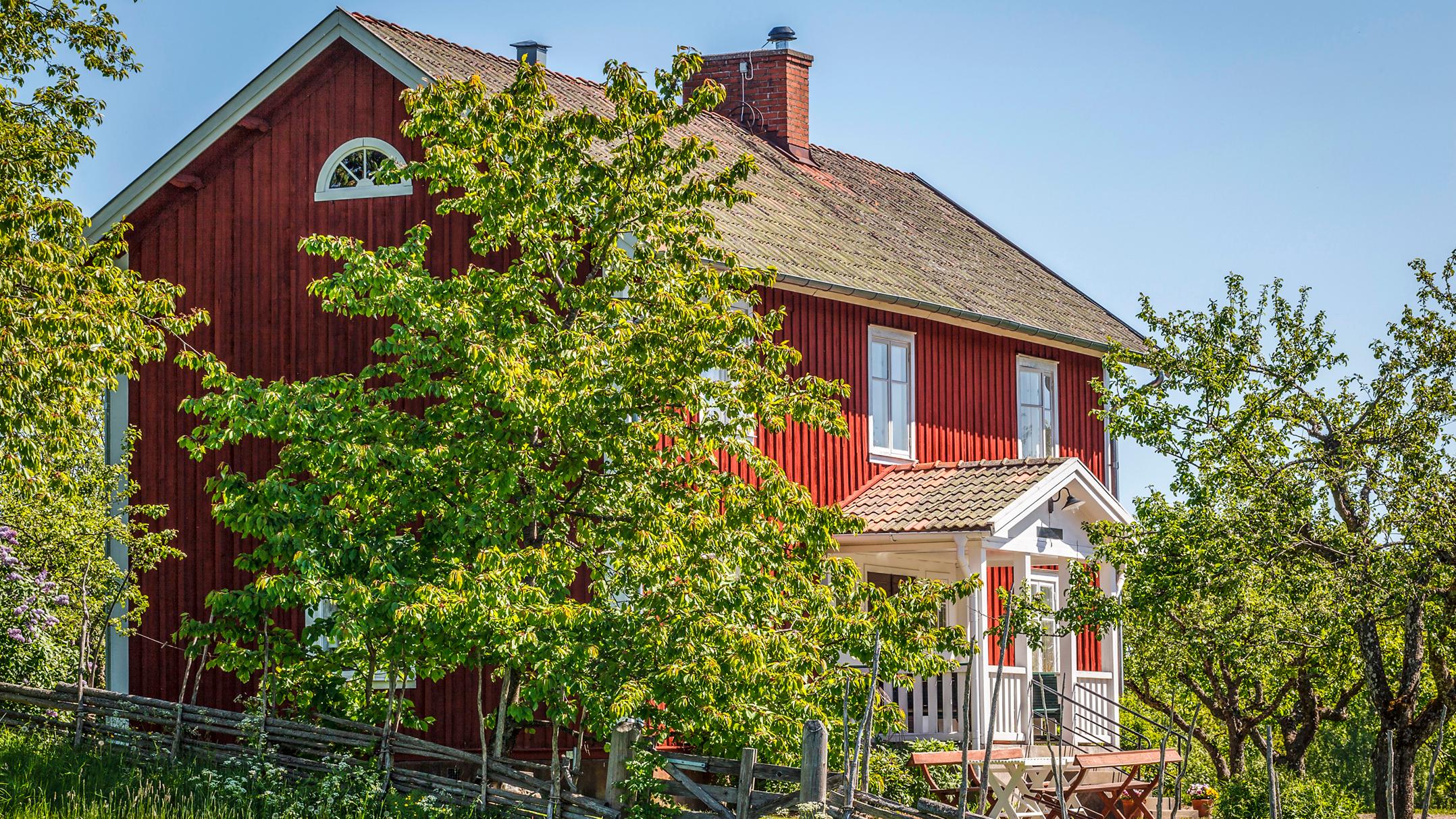 A red house with a white porch, surrounded by green trees, under a blue sky.