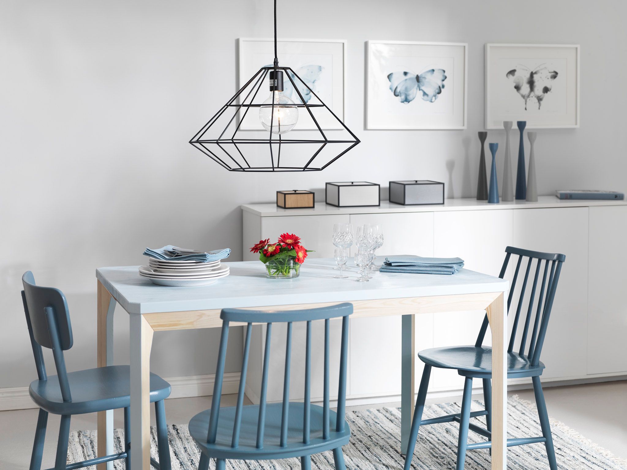 A dining room with a light blue table, three blue chairs, a black geometric pendant light, and three framed butterfly prints on the wall.