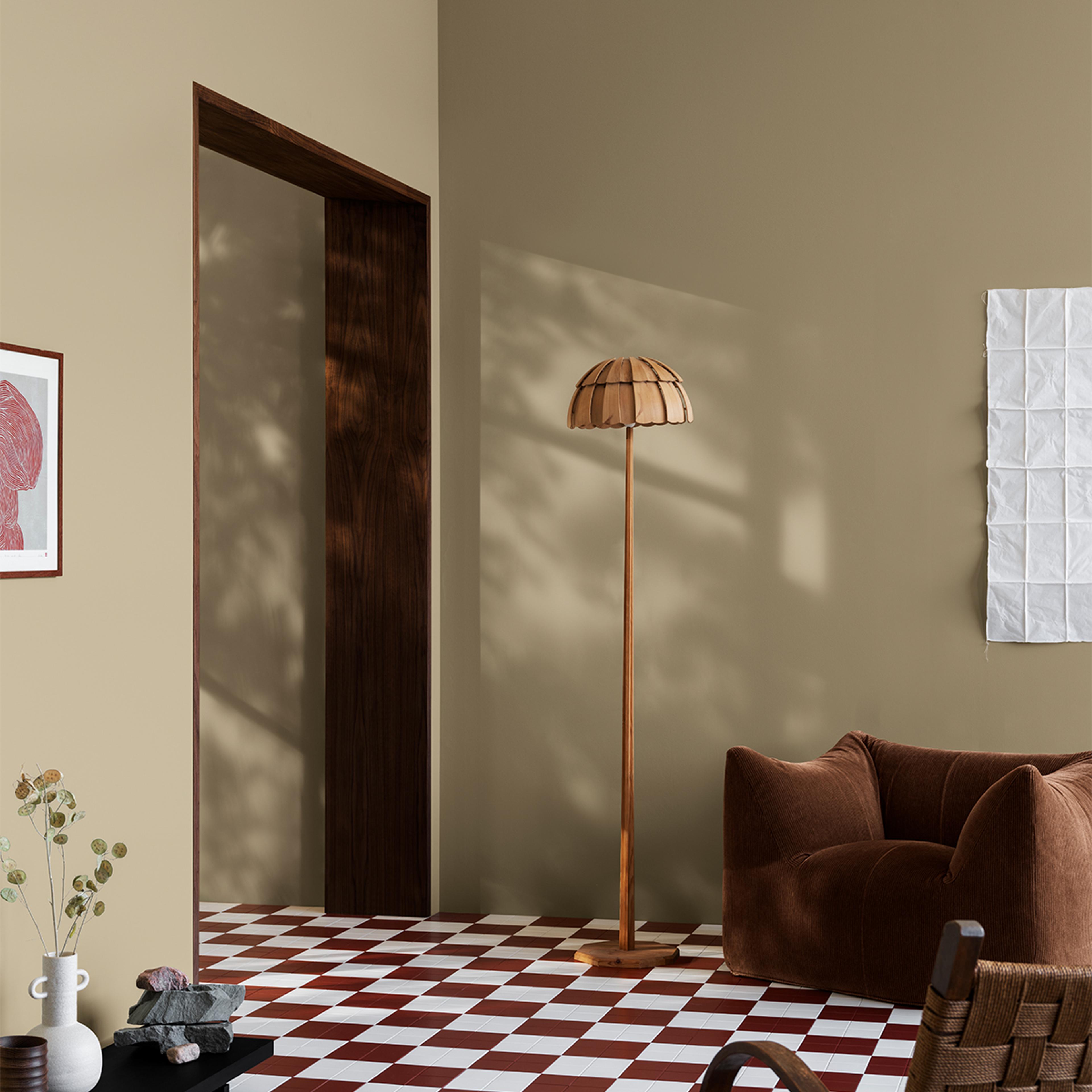 An inviting, earthy-toned room with a red and white checkered floor, a wooden floor lamp, a plush brown armchair, and sunlight casting tree shadows on the walls.