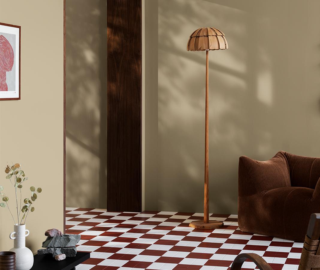 An inviting, earthy-toned room with a red and white checkered floor, a wooden floor lamp, a plush brown armchair, and sunlight casting tree shadows on the walls.
