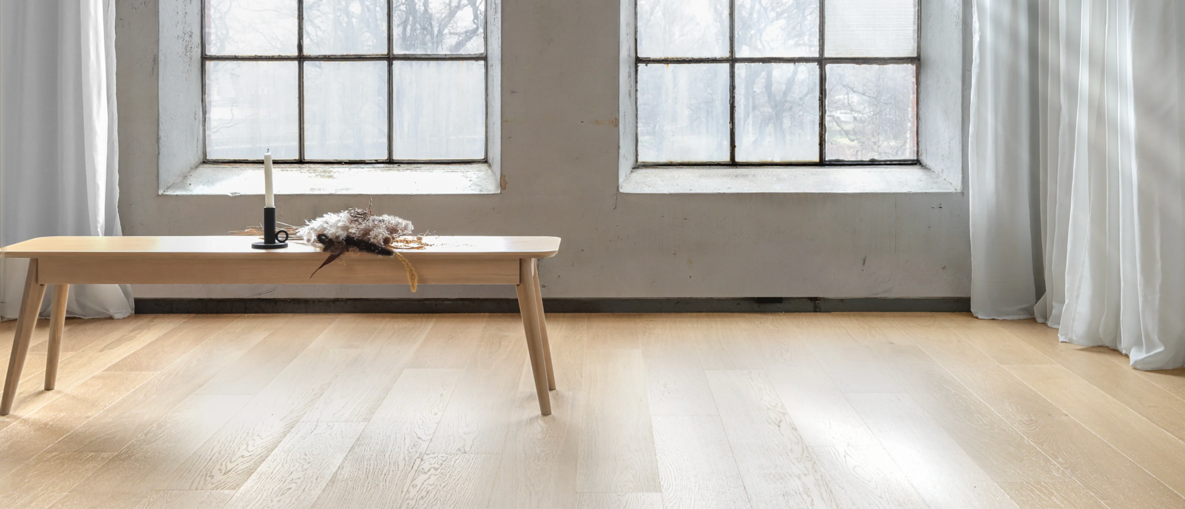 Light wood floor, two industrial-style windows, and a wooden bench with a candle and feathers.