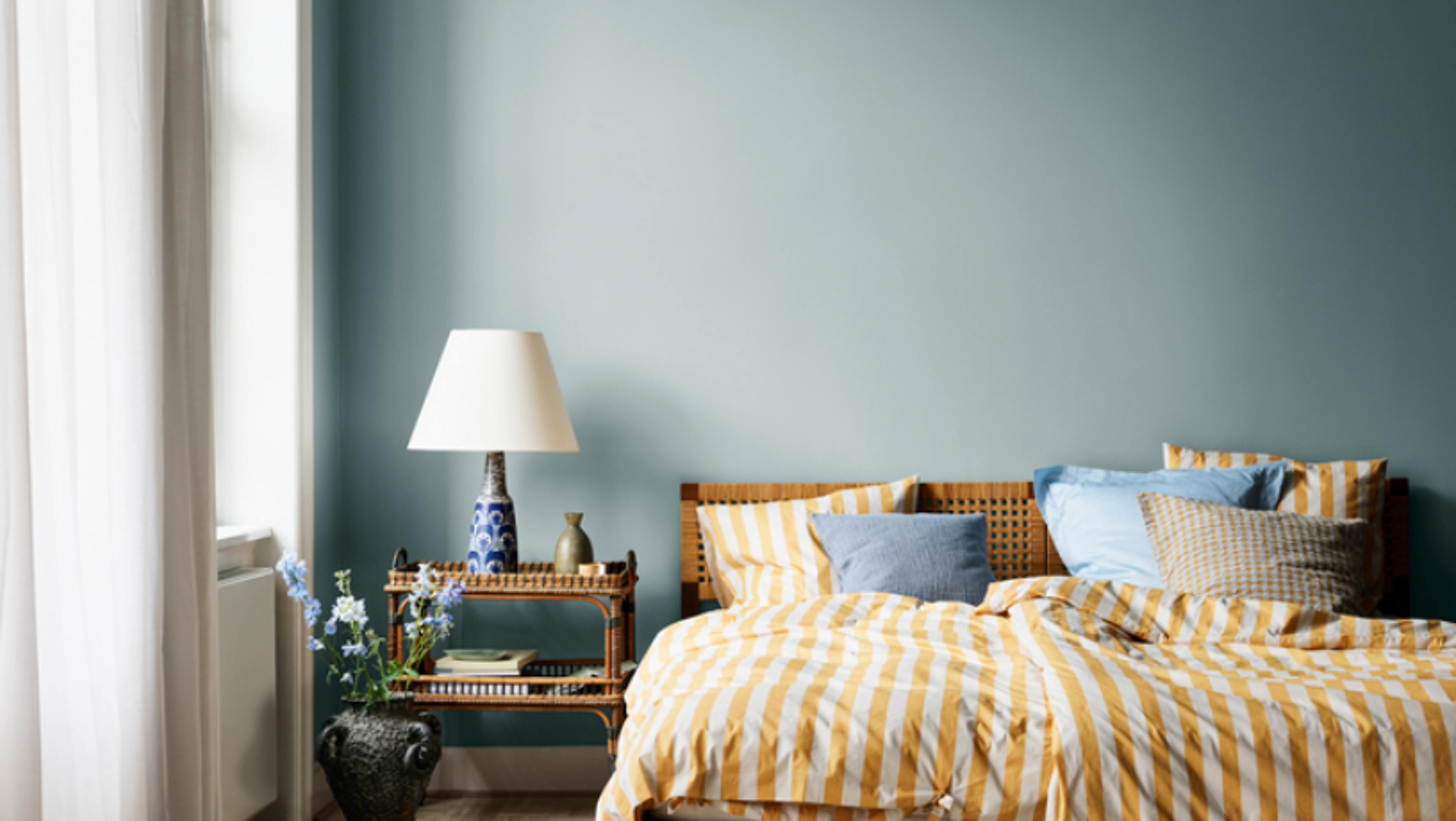 A bedroom with light blue walls, a bed with yellow and white striped bedding, and a wicker nightstand.