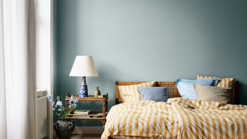 A bedroom with light blue walls, a bed with yellow and white striped bedding, and a wicker nightstand.