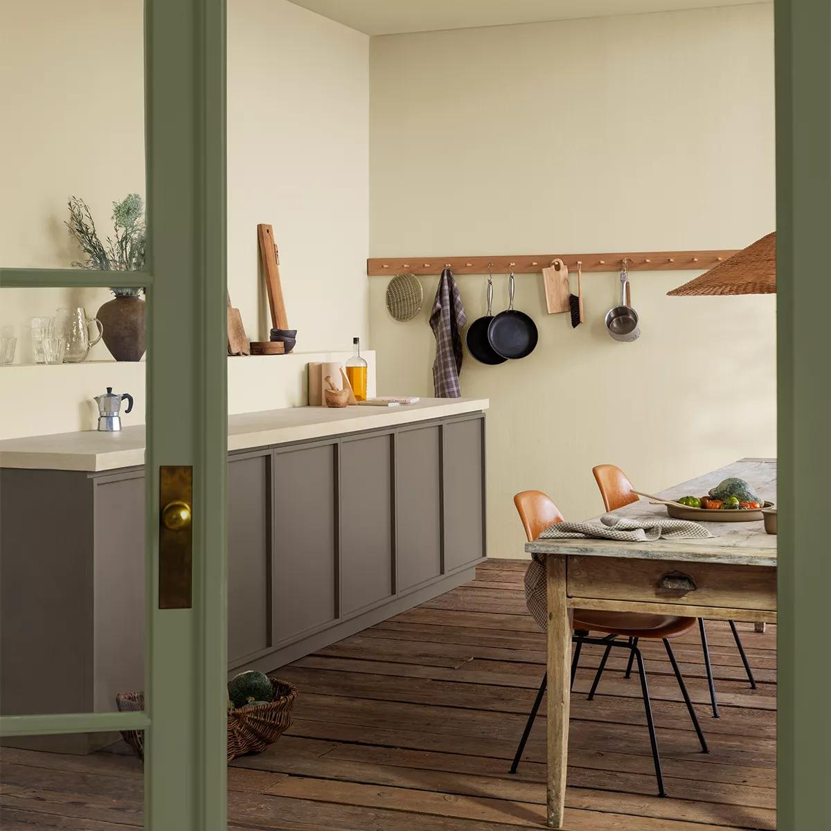 A rustic kitchen with cream walls, taupe cabinets, a peg rail, and a dining table, framed by a green door.