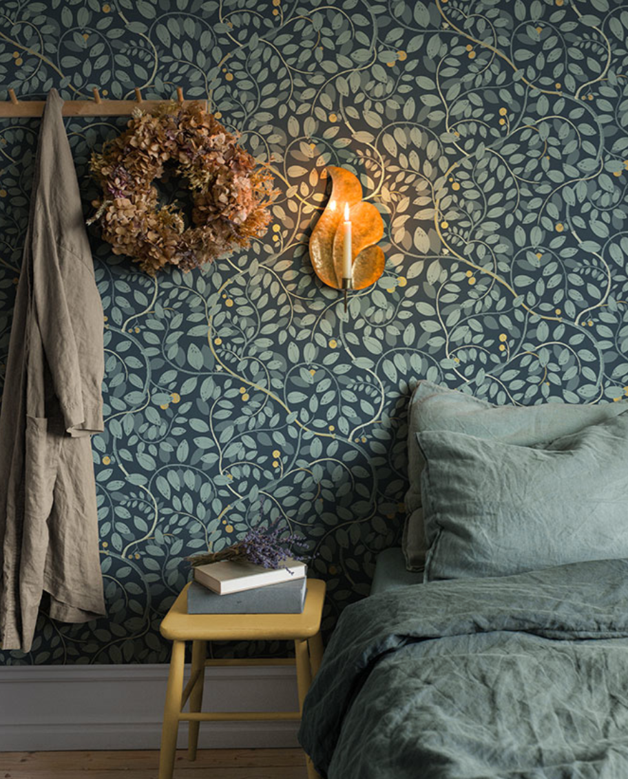 A cozy bedroom with dark teal leaf-patterned wallpaper, a lit candle sconce, a dried floral wreath, and a bed with green linen.