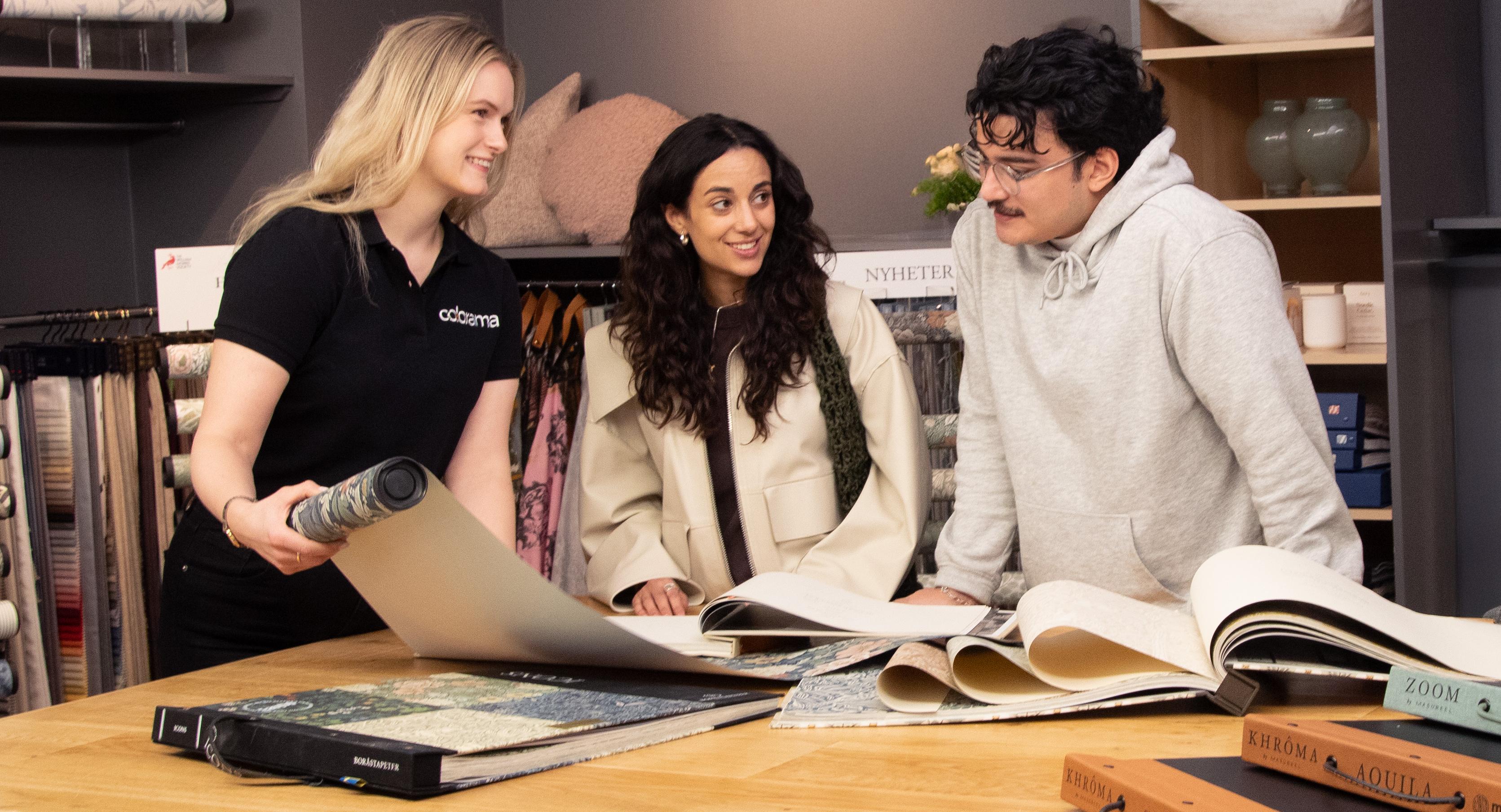 An employee assists two customers browsing wallpaper and fabric samples in a design store.