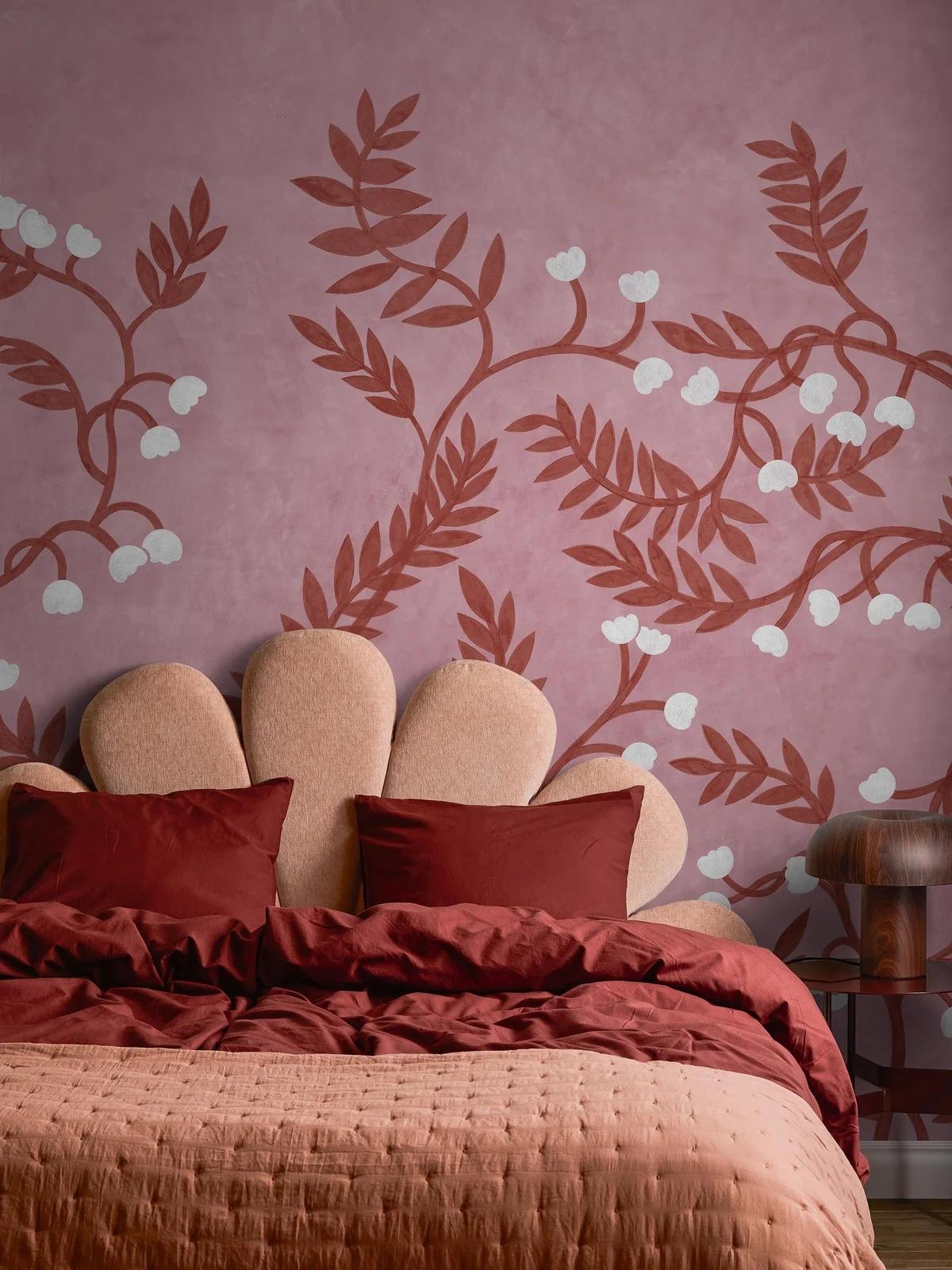 A bedroom with a pink floral wall mural above a bed with a scalloped headboard and red and pink bedding.