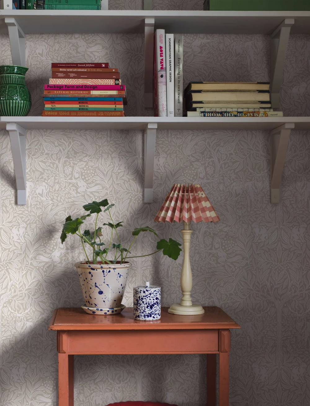 White shelves holding books and a green vase above an orange table with a potted plant, a speckled jar, and a lamp with a pink checkered shade, set against patterned wallpaper.