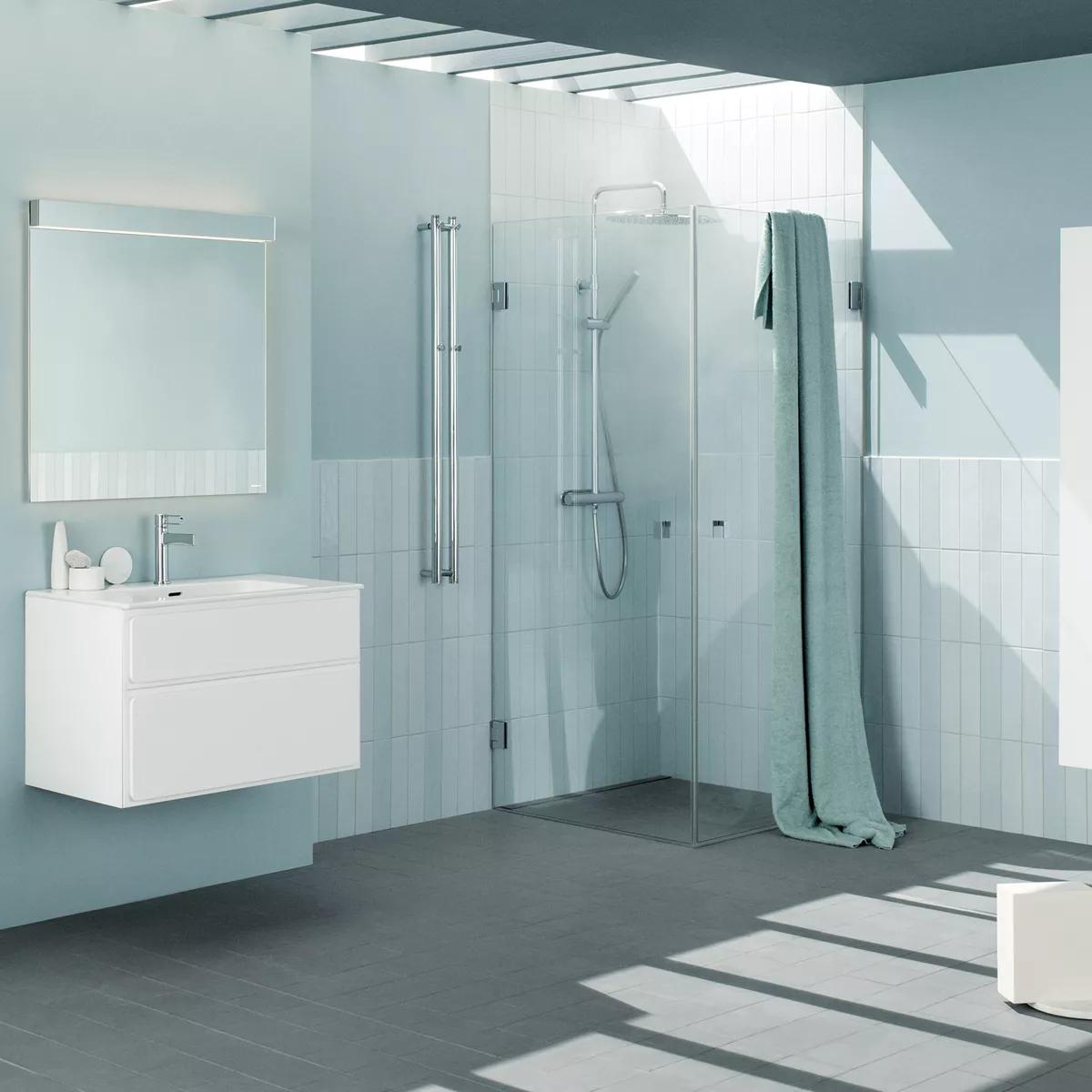 Modern light blue bathroom with a white floating vanity, glass-enclosed shower, green towel, and sunlight on a grey tiled floor.
