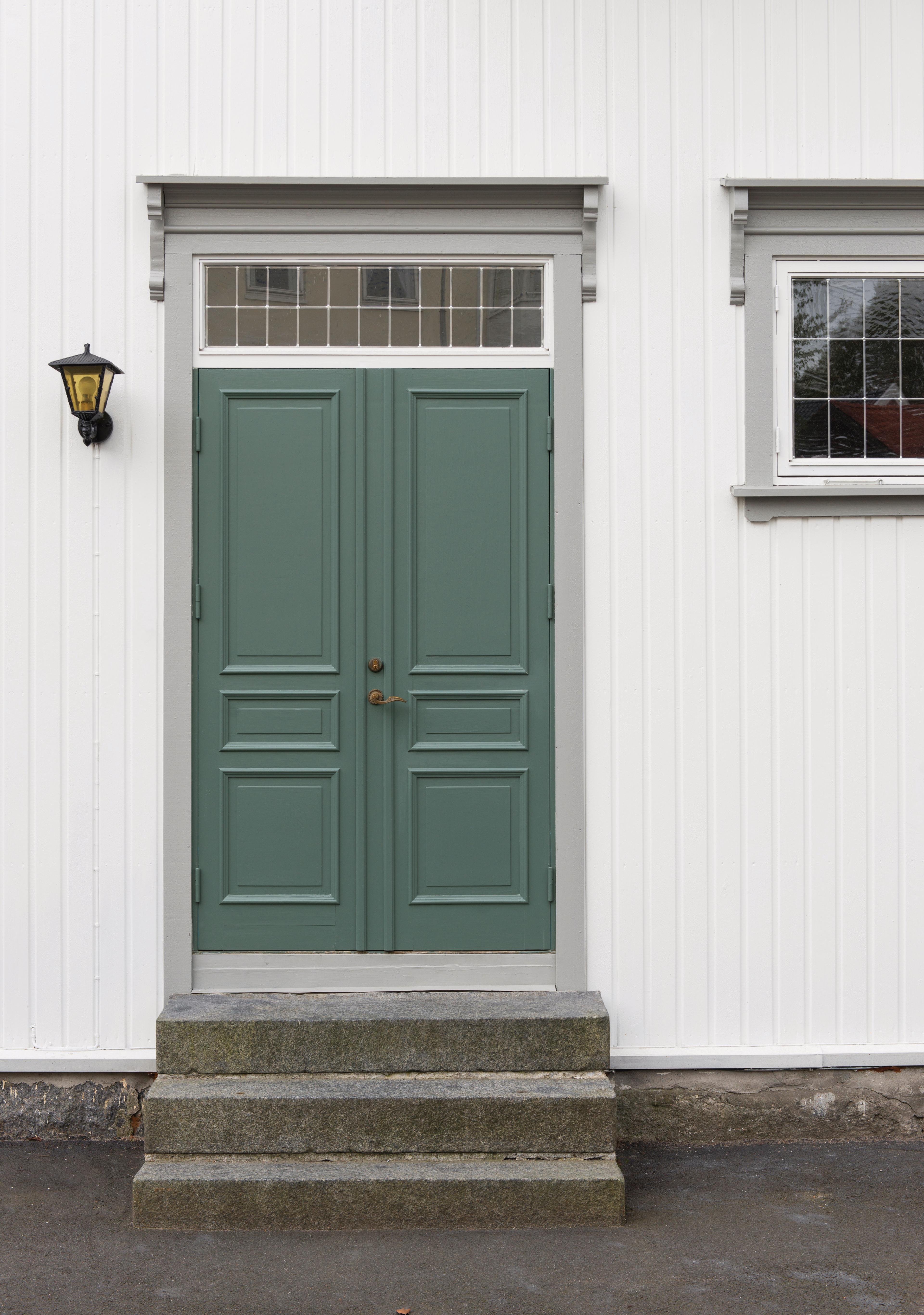A green double door with a transom window, three stone steps, a lantern, and a side window on a white building.