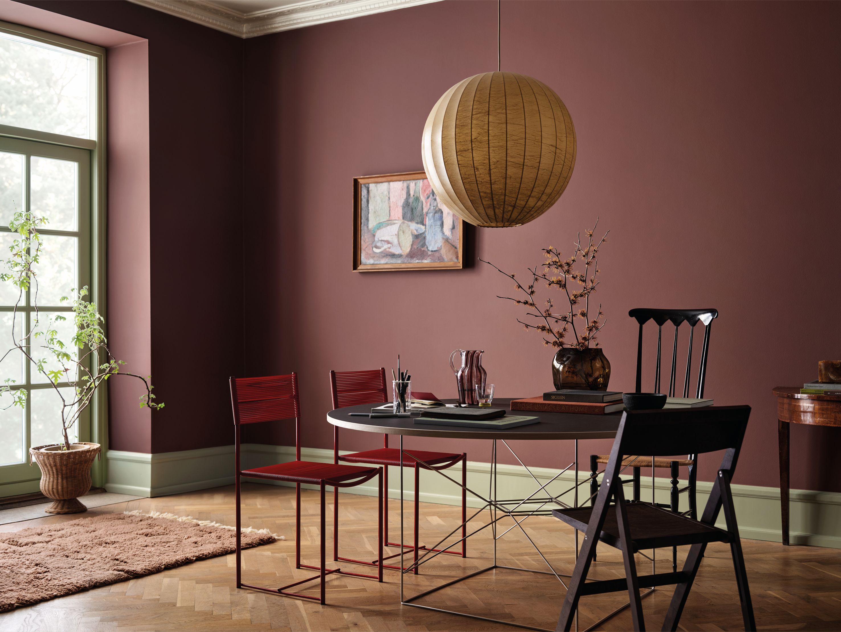 A modern dining room with dark rose walls, a spherical paper lantern, a round table, and mismatched chairs on a hardwood floor.