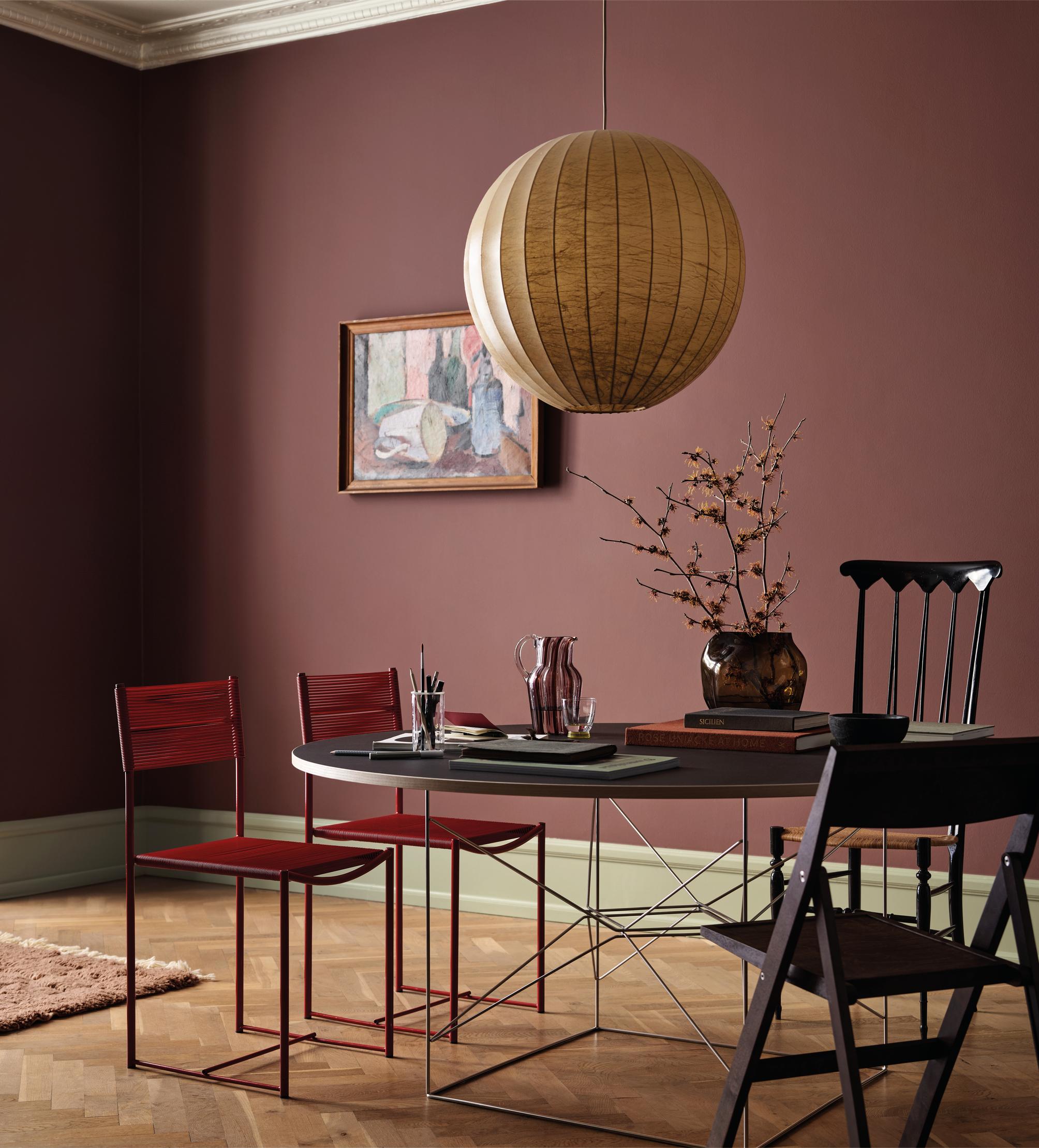 A modern dining room with dark rose walls, a spherical paper lantern, a round table, and mismatched chairs on a hardwood floor.