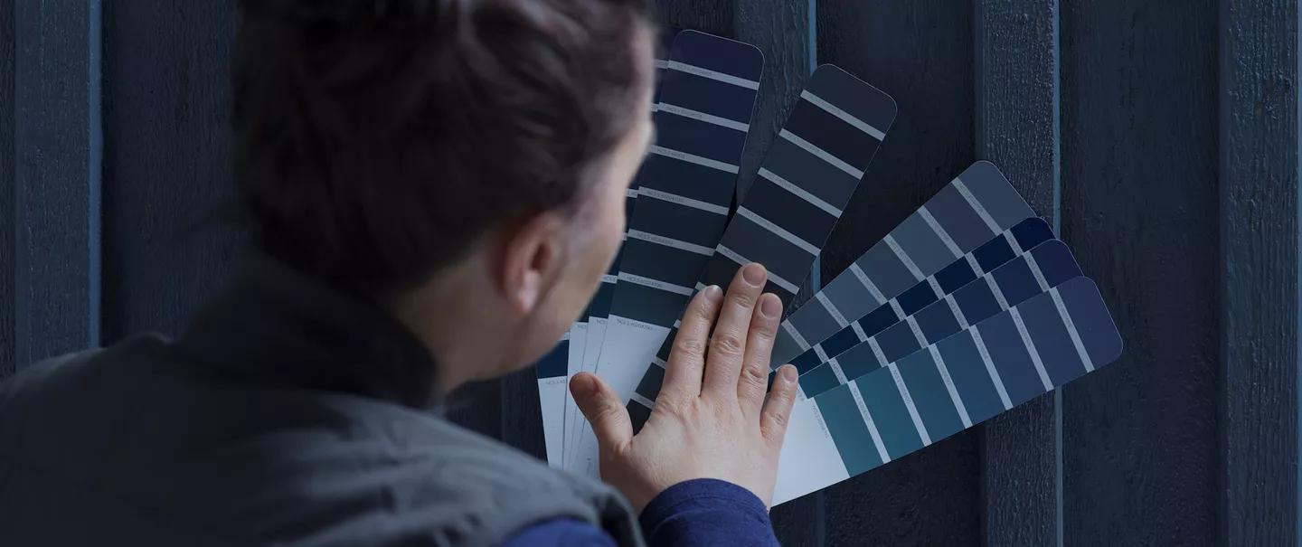 A person's hand holds blue and gray paint swatches against a dark blue wooden wall.