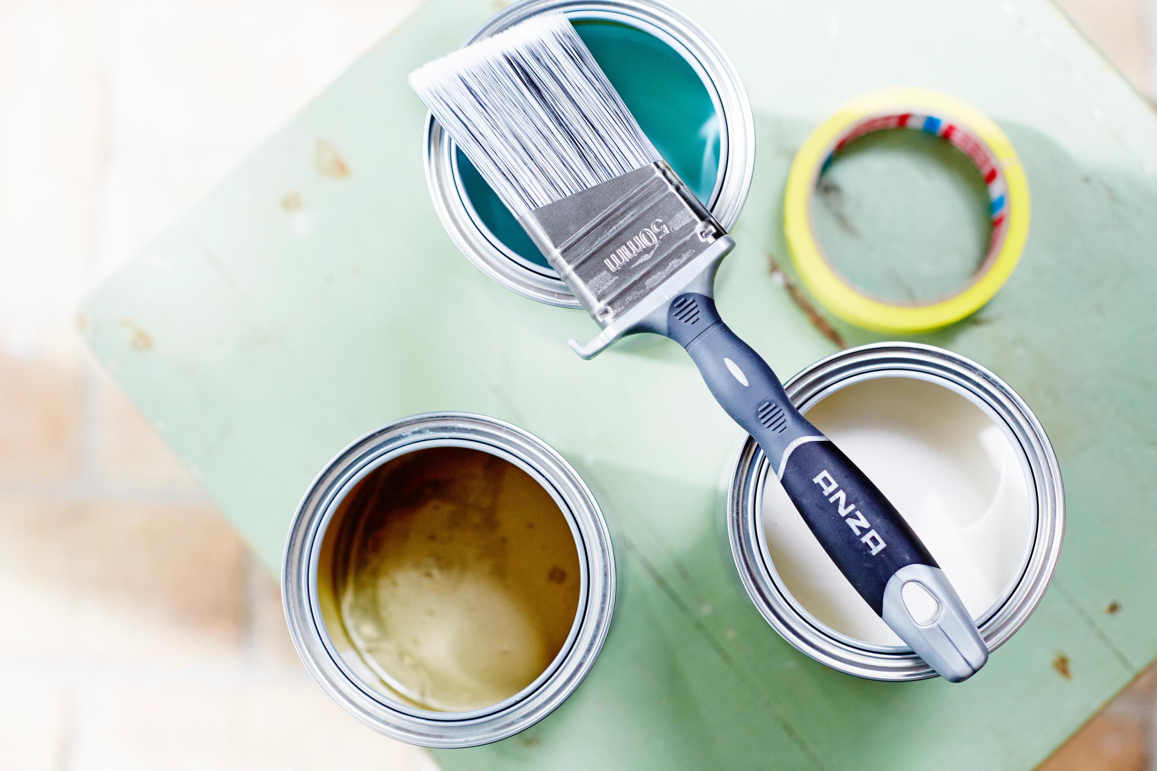 Open cans of teal, gold, and white paint, a brush, and masking tape on a green surface.