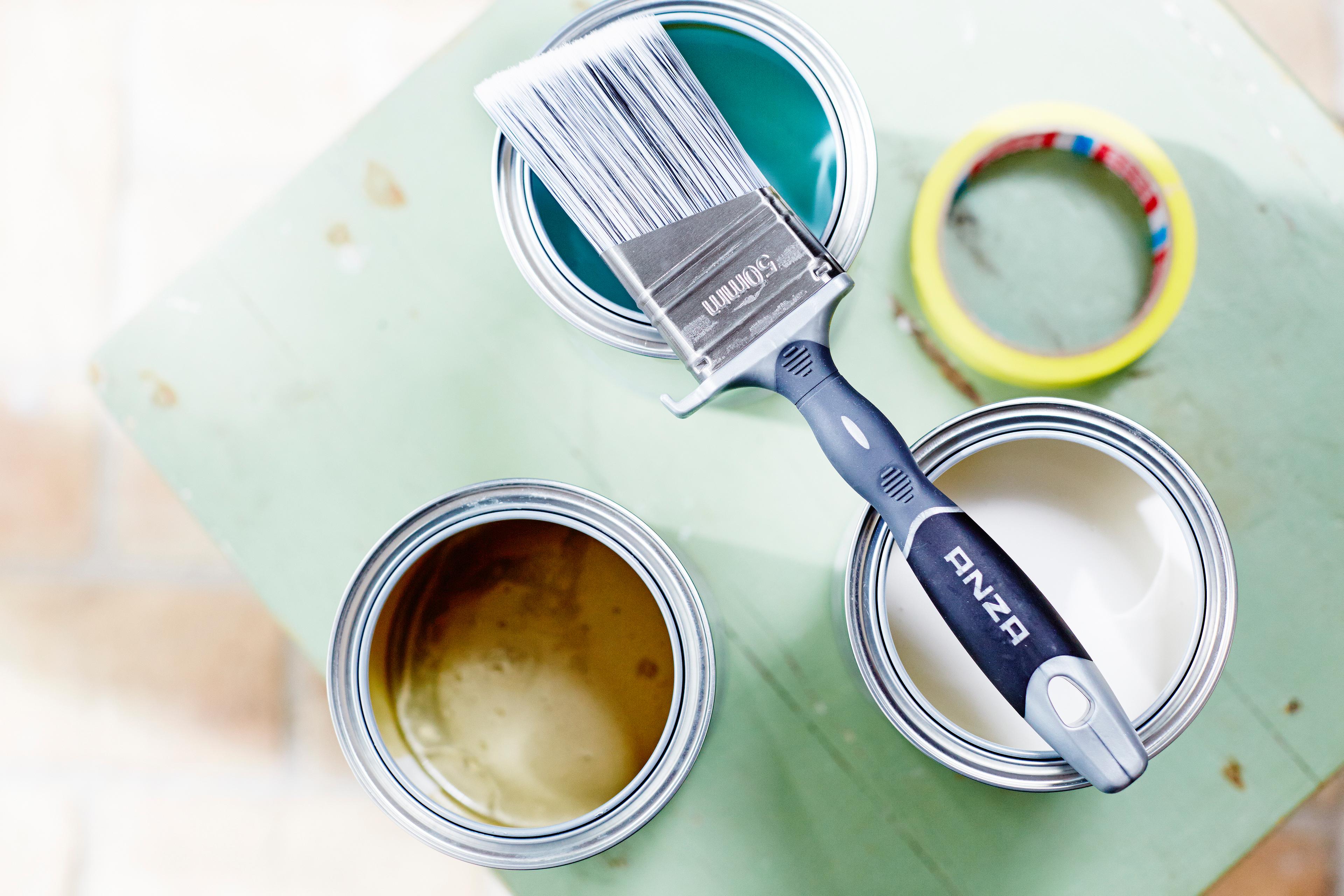 Three open paint cans (teal, gold, white) with a paintbrush and masking tape on a green surface.